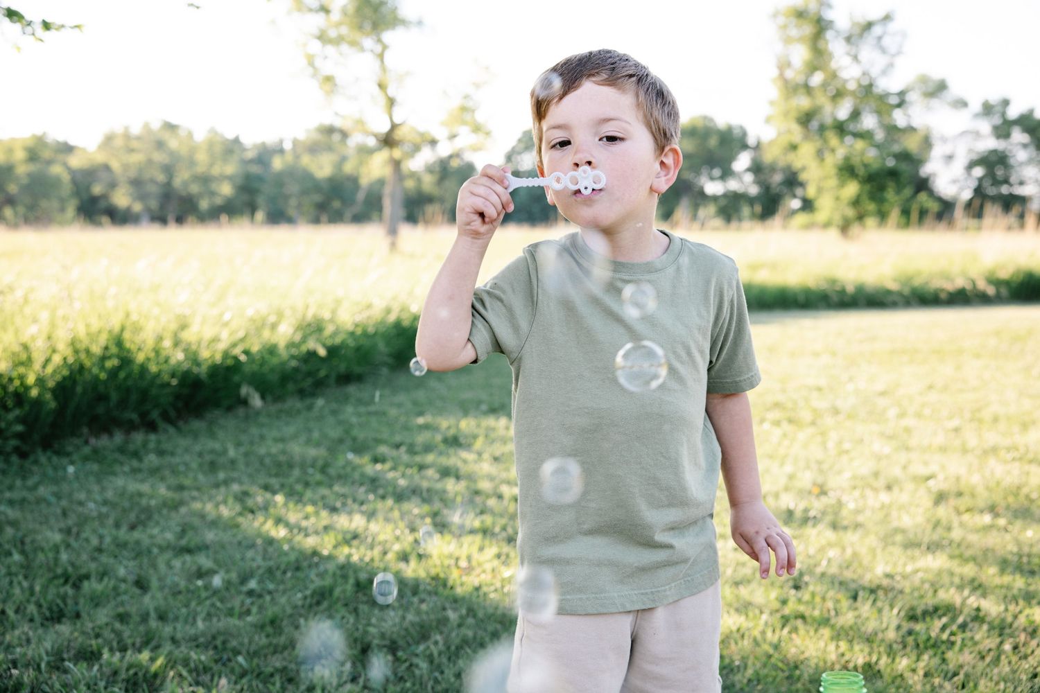 Young boy in green t-shirt blowing soap bubbles outdoors in a sunny green park.