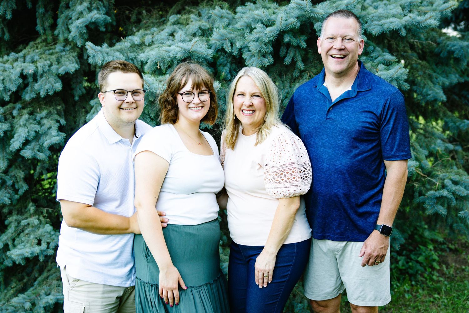 A smiling family of four poses outdoors near evergreen trees on a sunny day.