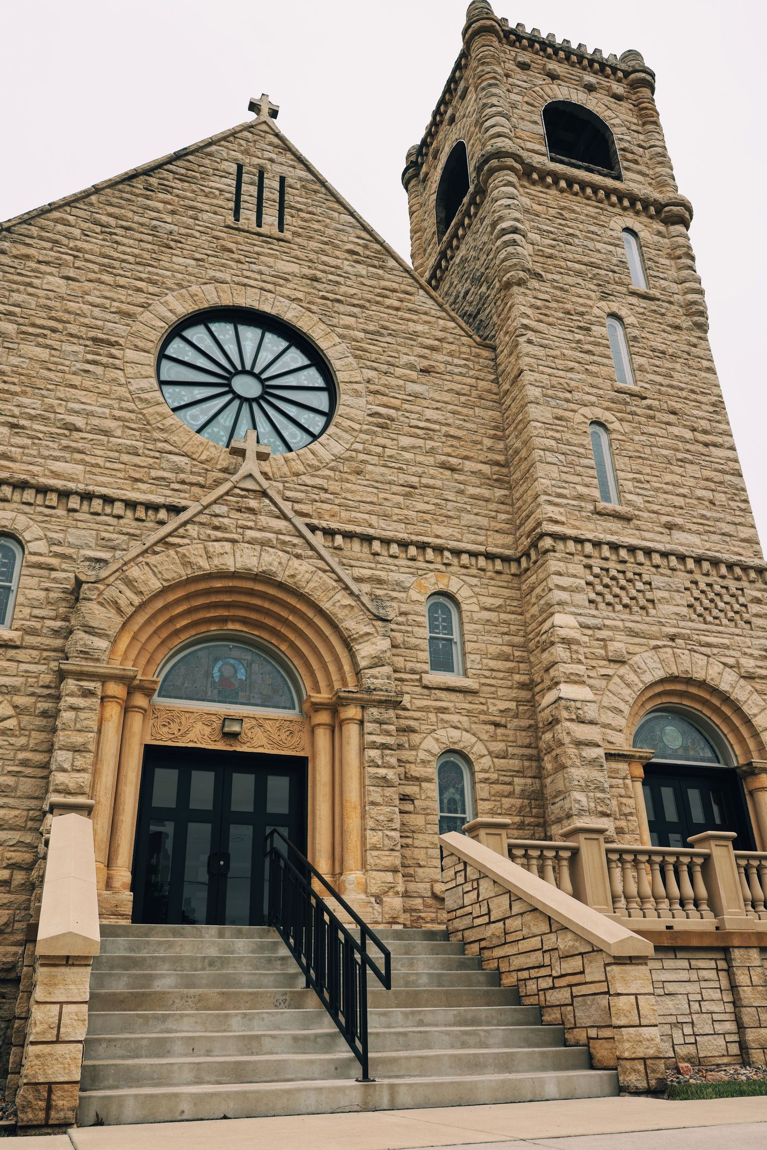 Historic stone Catholic church exterior with rose window, arched entrance, bell tower, and grand staircase under overcast sky.