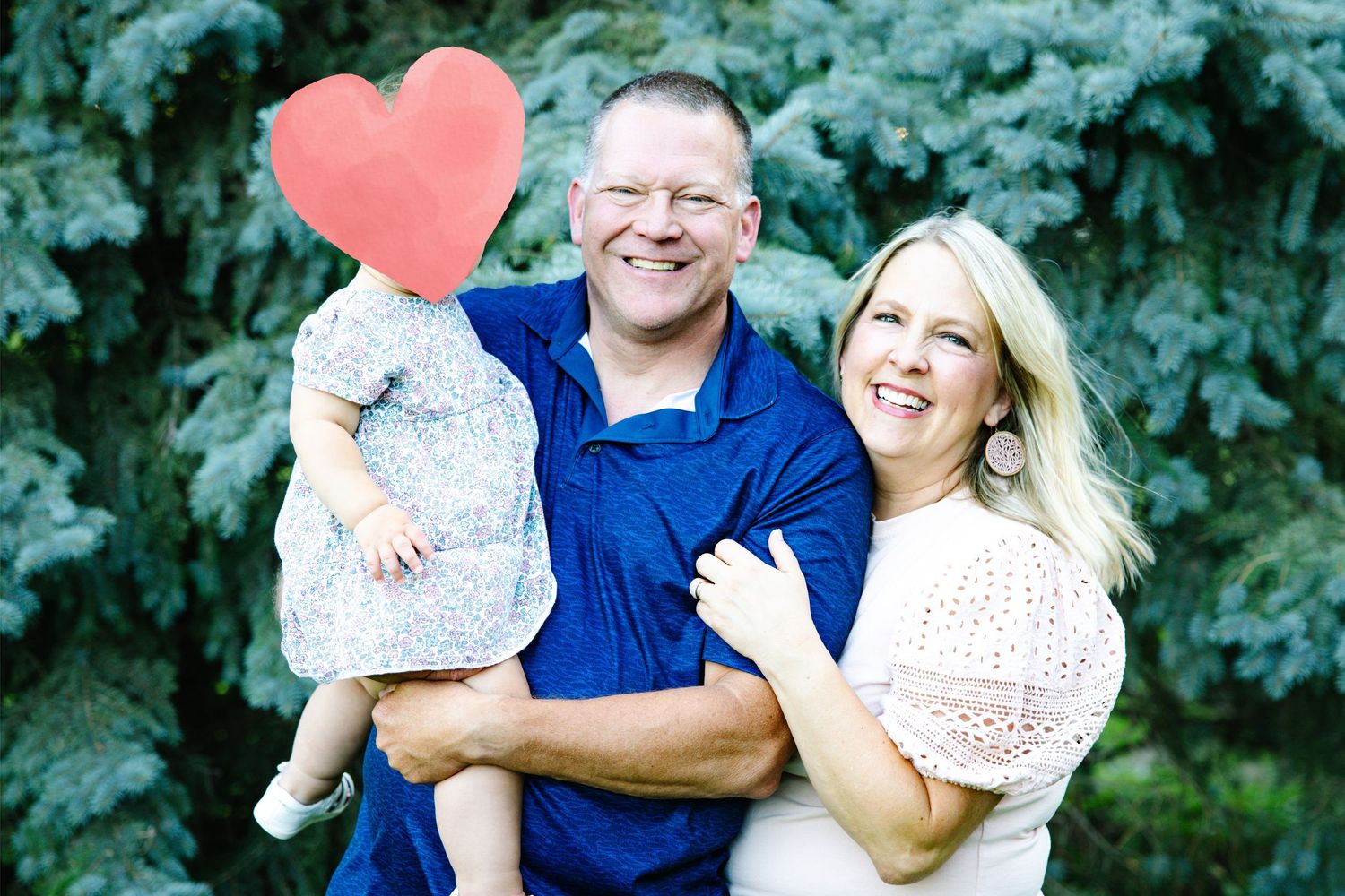 A smiling couple poses outdoors holding a child with a red heart covering their face, with blue spruce trees behind them.