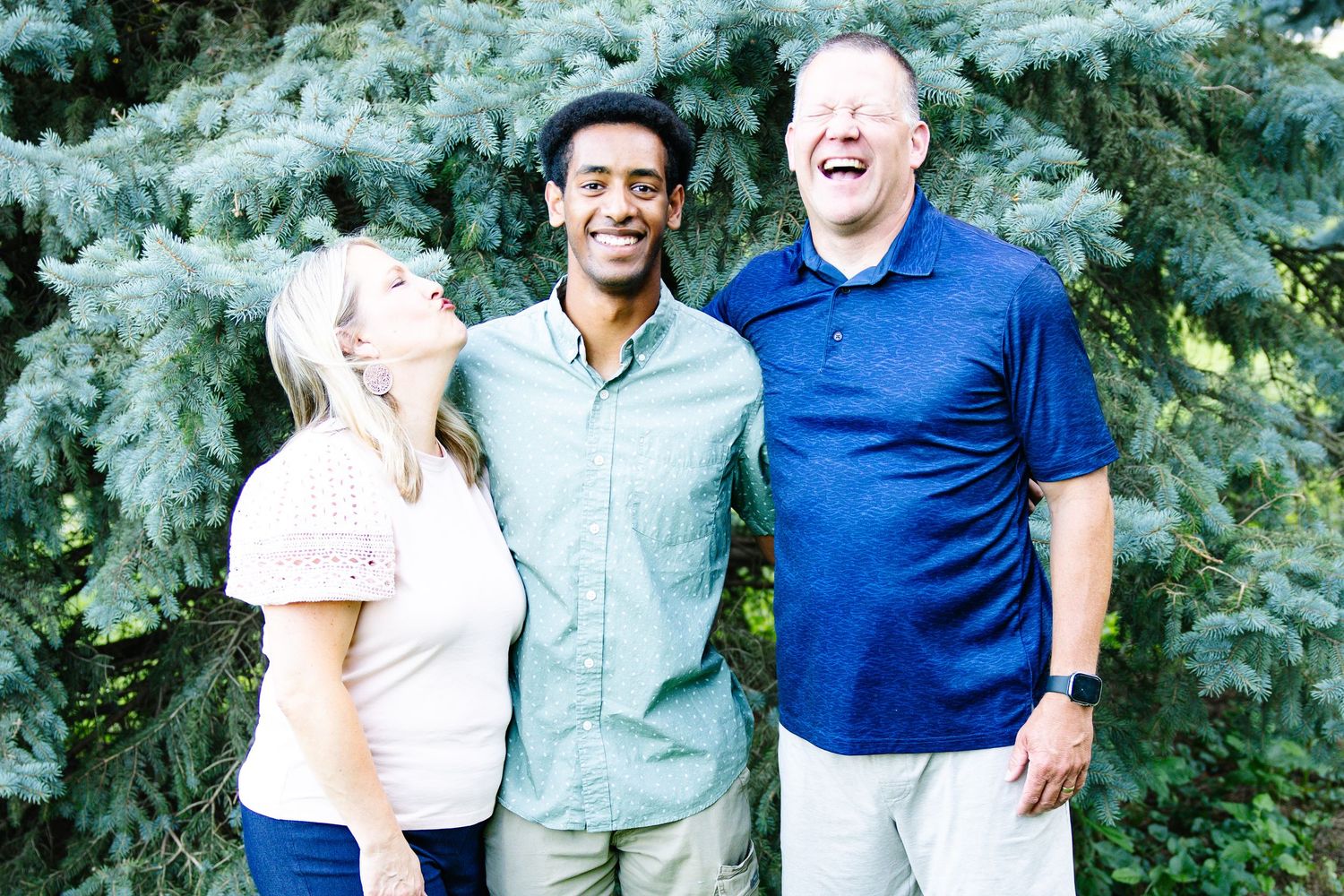 Three people smiling and laughing together outdoors in front of a lush blue spruce tree on a sunny day.