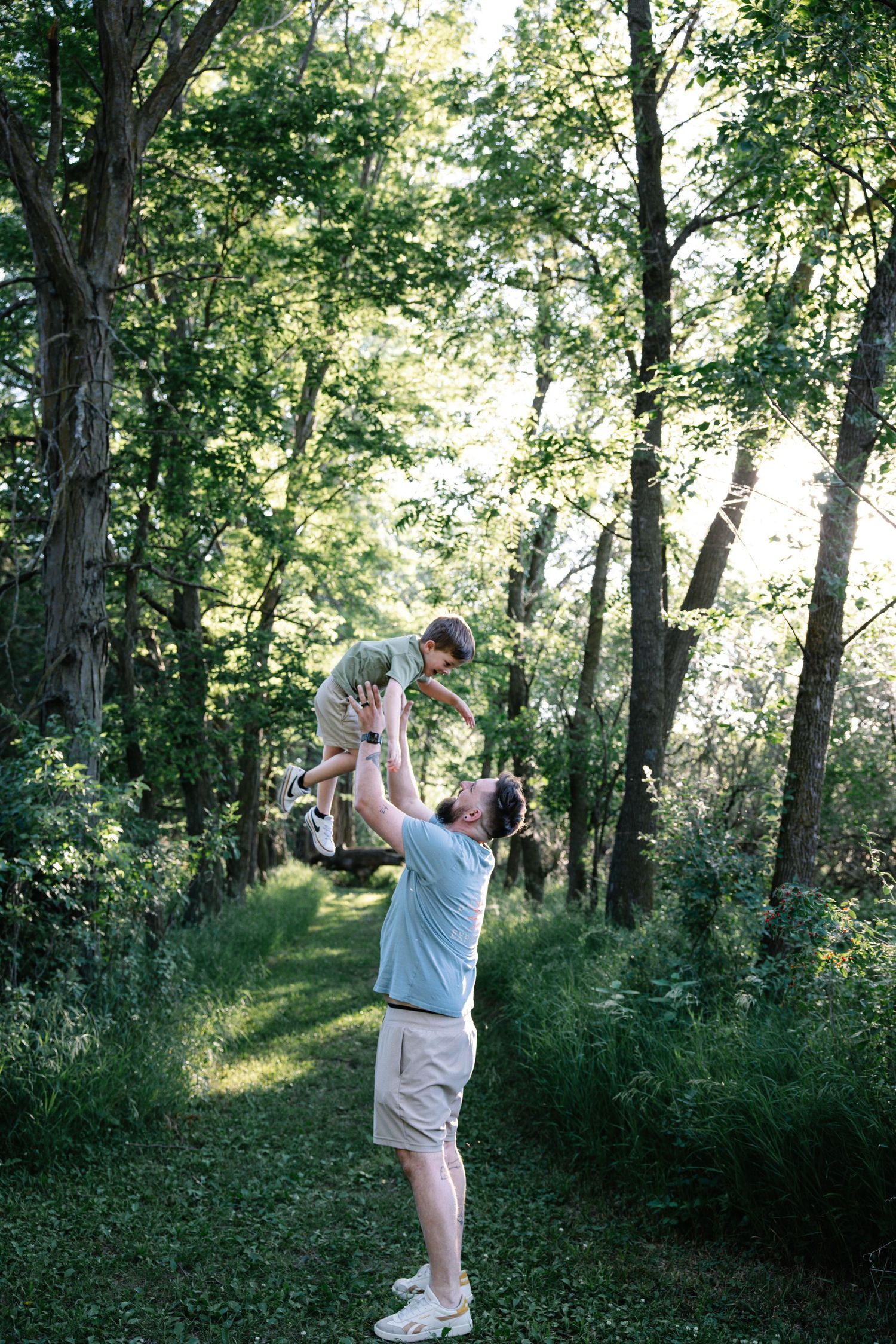 Father joyfully lifting young child overhead on a sunny forest path surrounded by lush green trees.