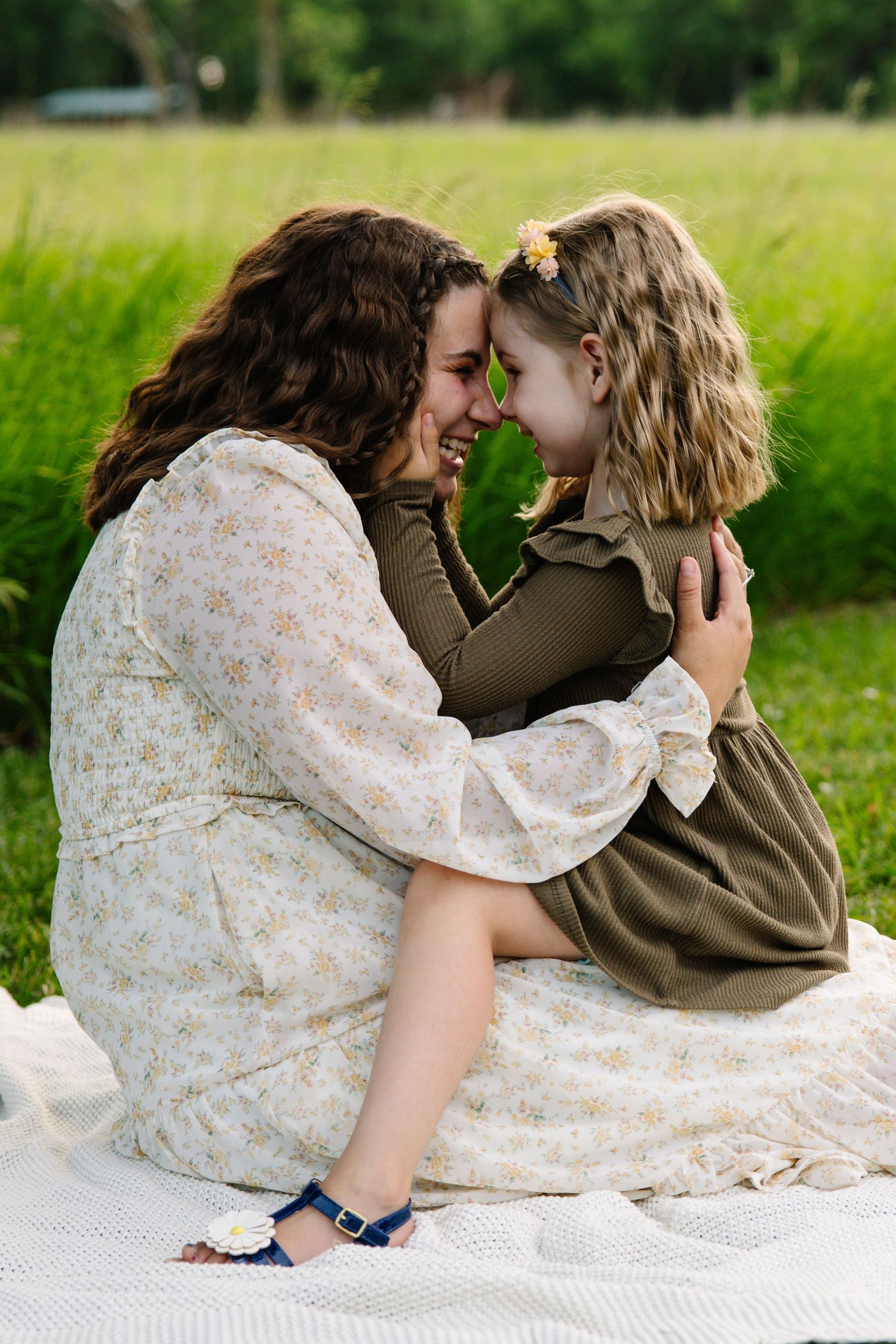 A mother and daughter touch foreheads and laugh while sitting on a blanket outside.