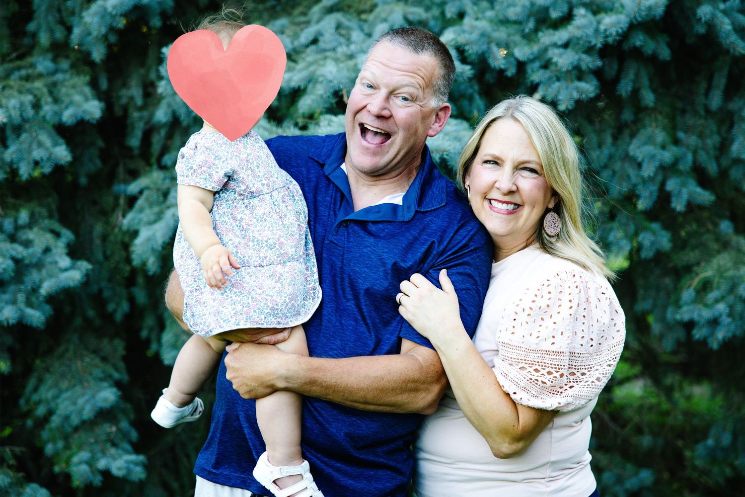 Happy family portrait with smiling man holding toddler and blonde woman posing outdoors near blue spruce trees.