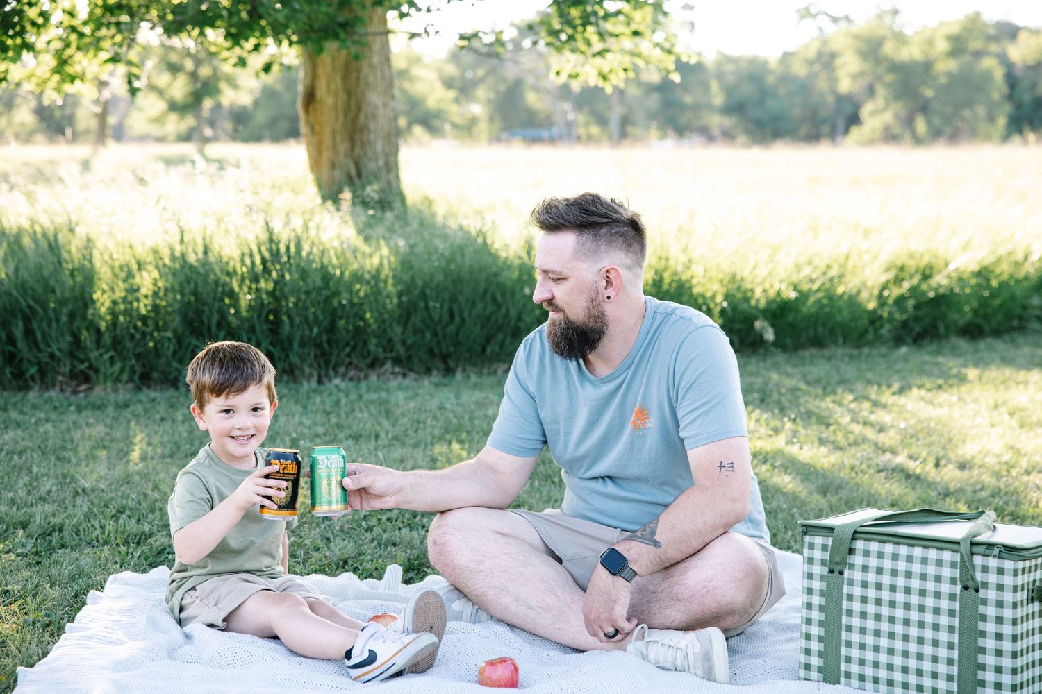 Father and young son share a drink together on a picnic blanket in a sunny green park.