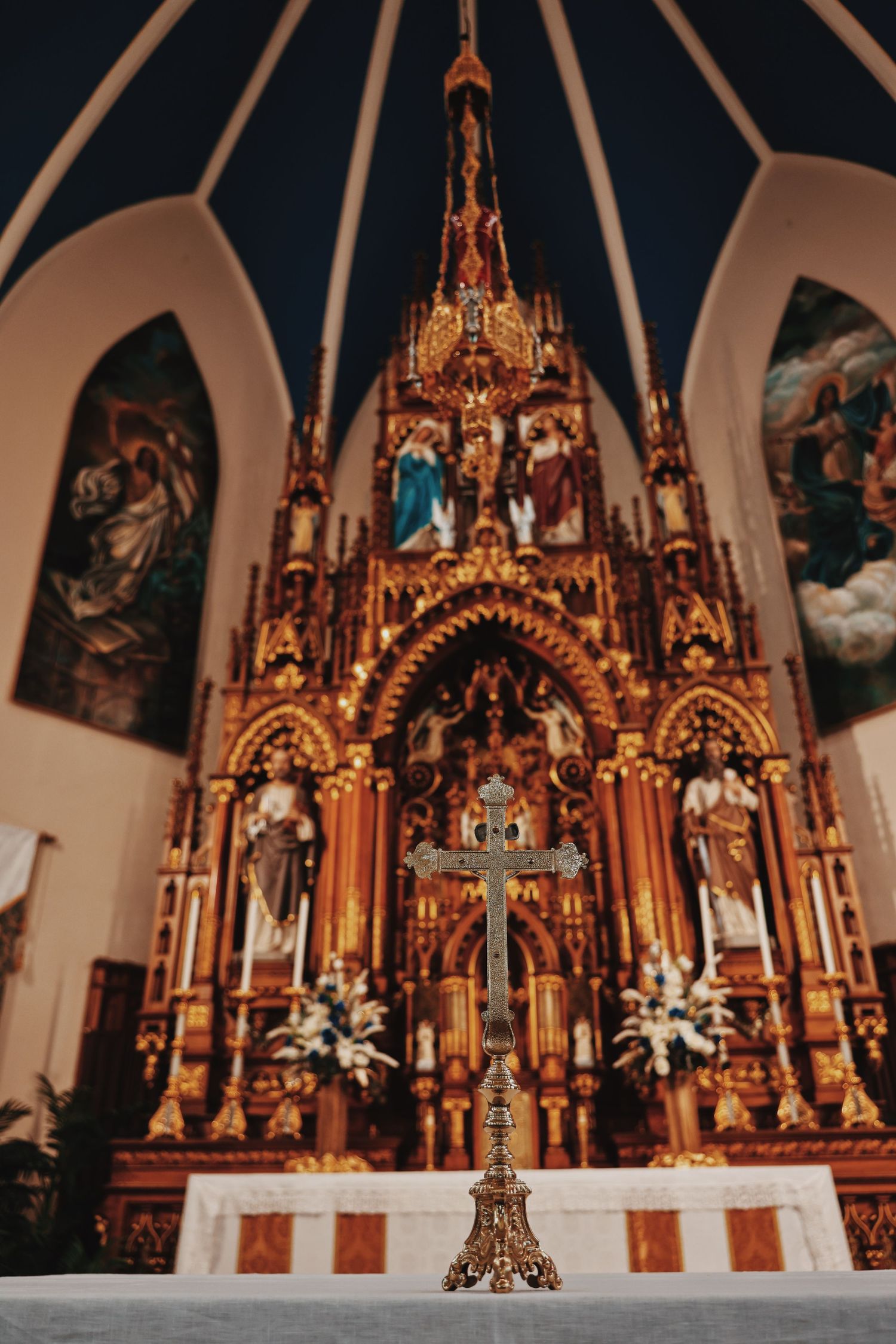 Ornate golden Catholic altar with intricate Gothic details, religious statues, paintings, and a decorative cross in foreground.