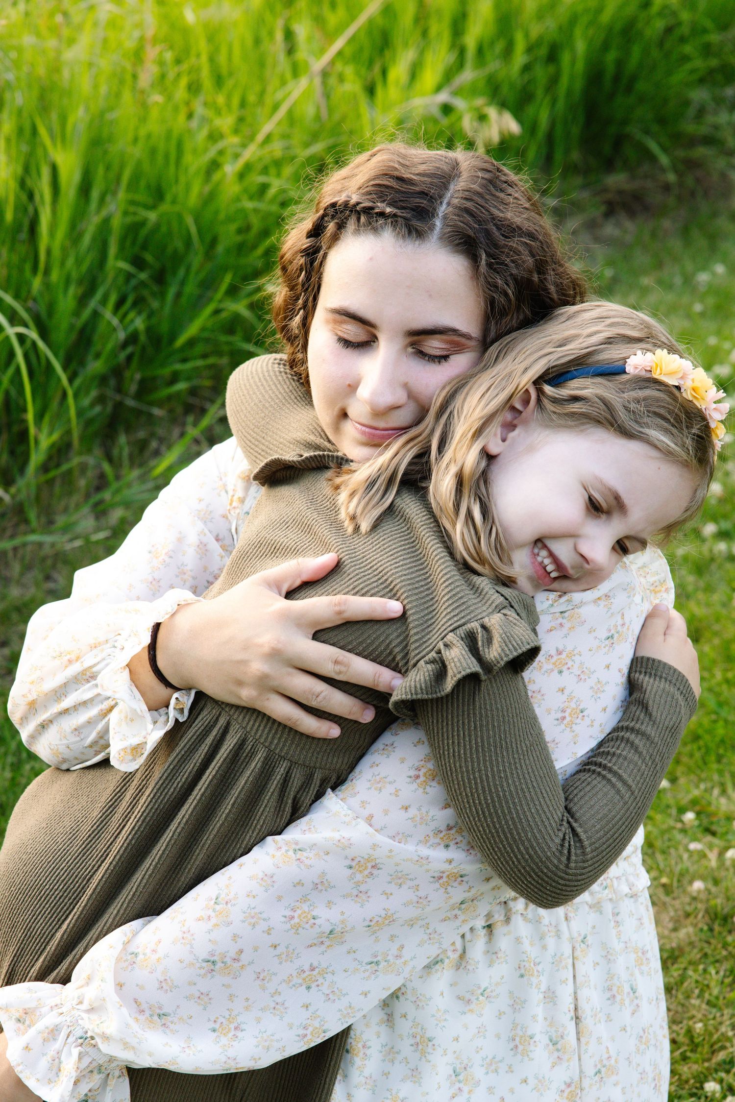 A teen girl warmly hugs a smiling young girl with a flower headband in a green meadow.