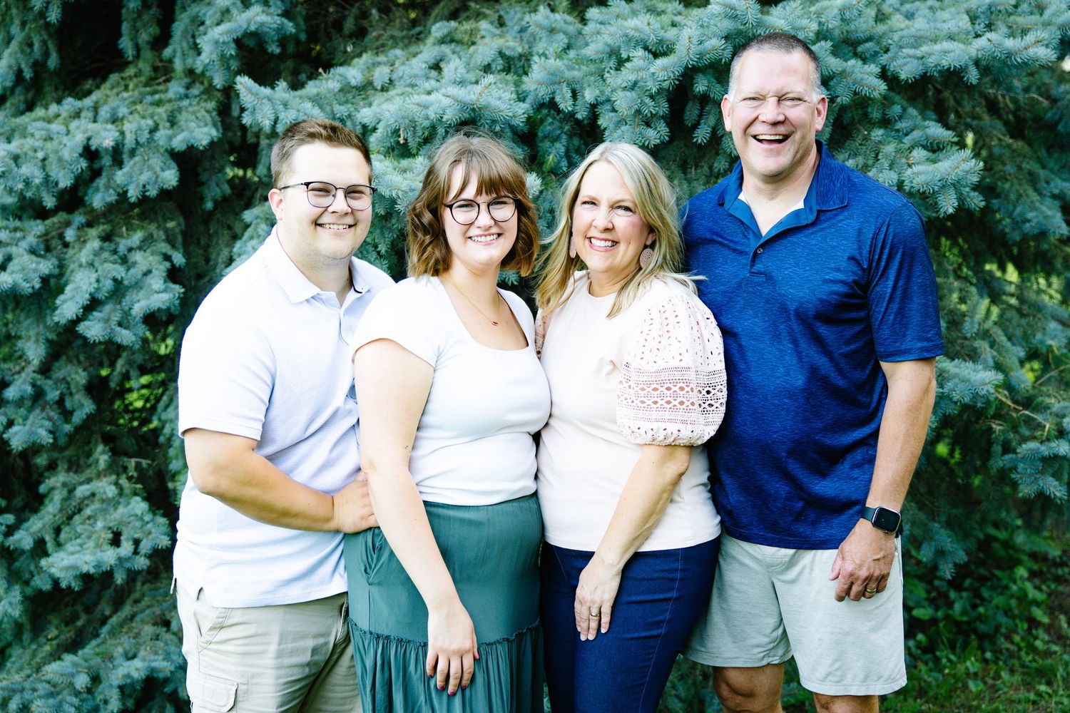 Four smiling family members pose together outdoors in front of lush green trees on a sunny day.