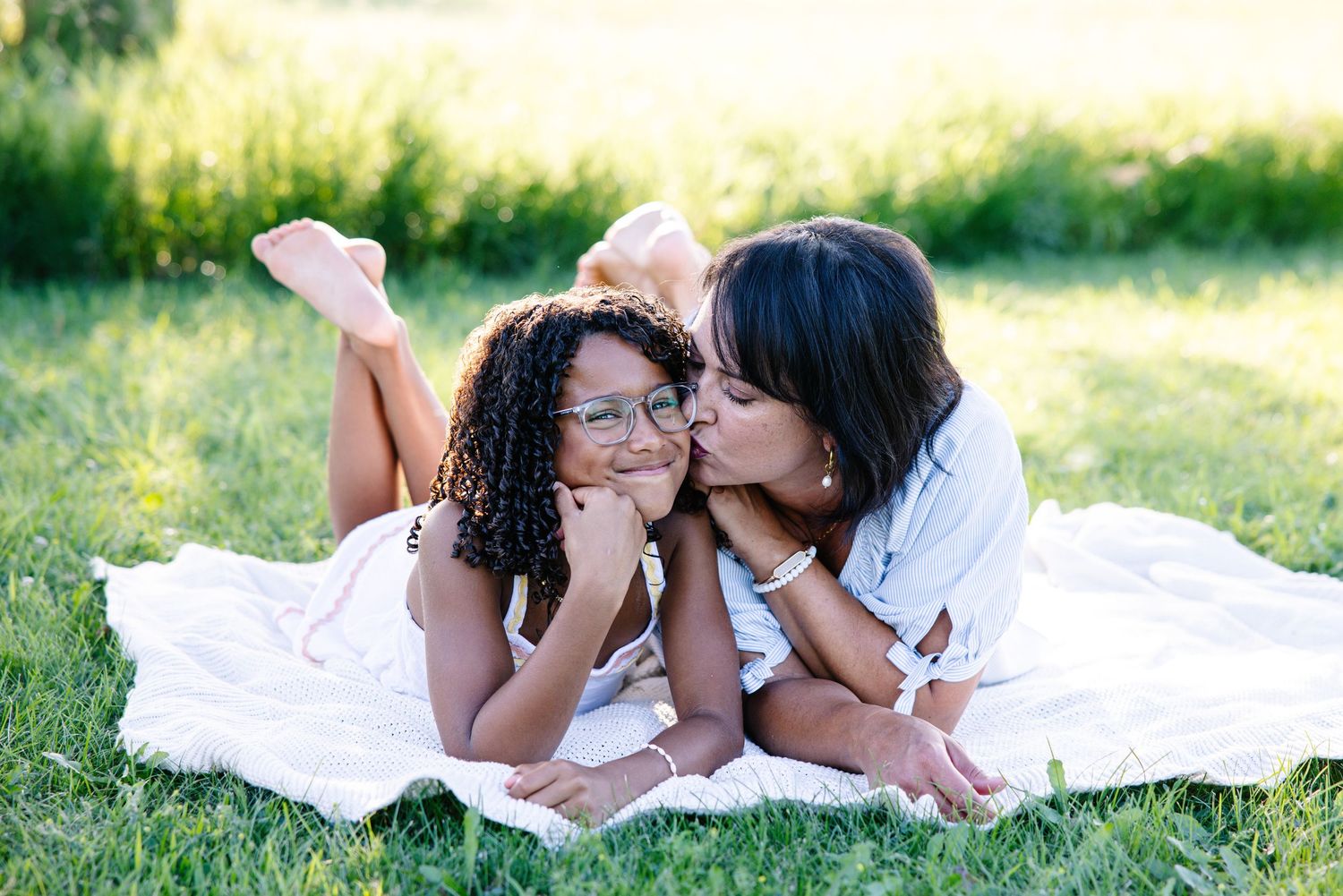 A mother kisses her daughter on the cheek as they lie on a blanket in a sunny green park.