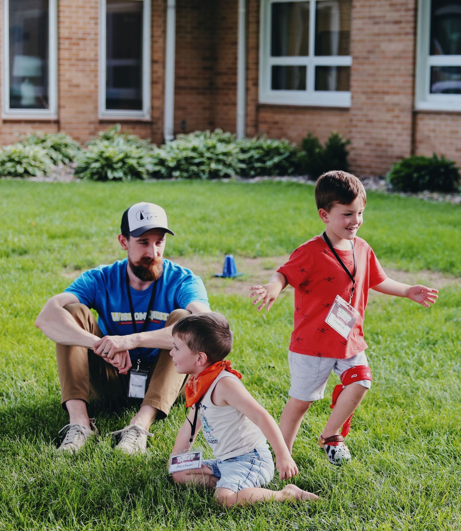 A bearded man in a blue shirt and cap sits on grass watching two young boys play outdoors near a brick building.