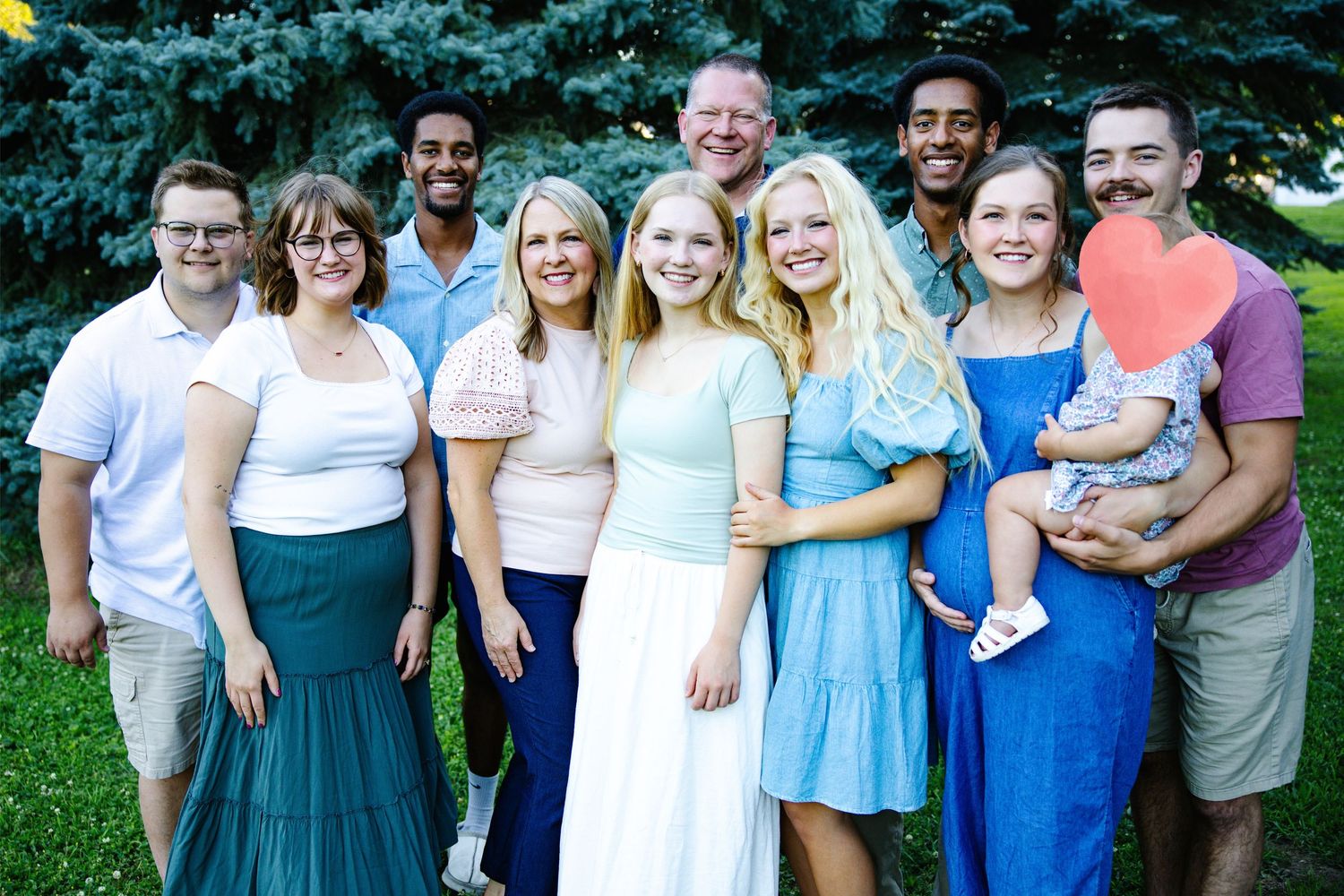 Large family group smiling outdoors near green trees, dressed in coordinating blue and white outfits for a family portrait.