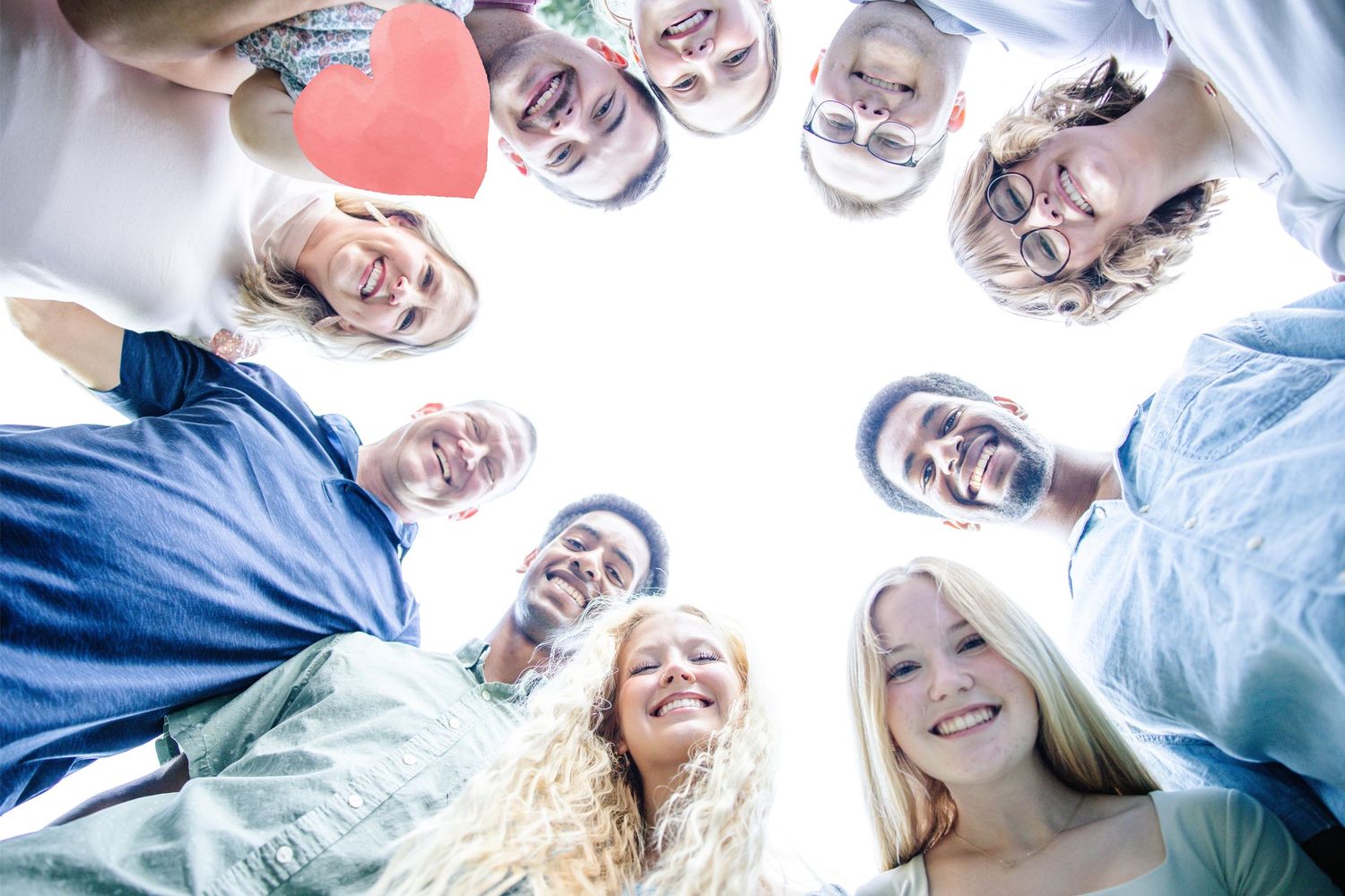 Diverse group of smiling young people forming a circle huddle, viewed from below against bright sky.