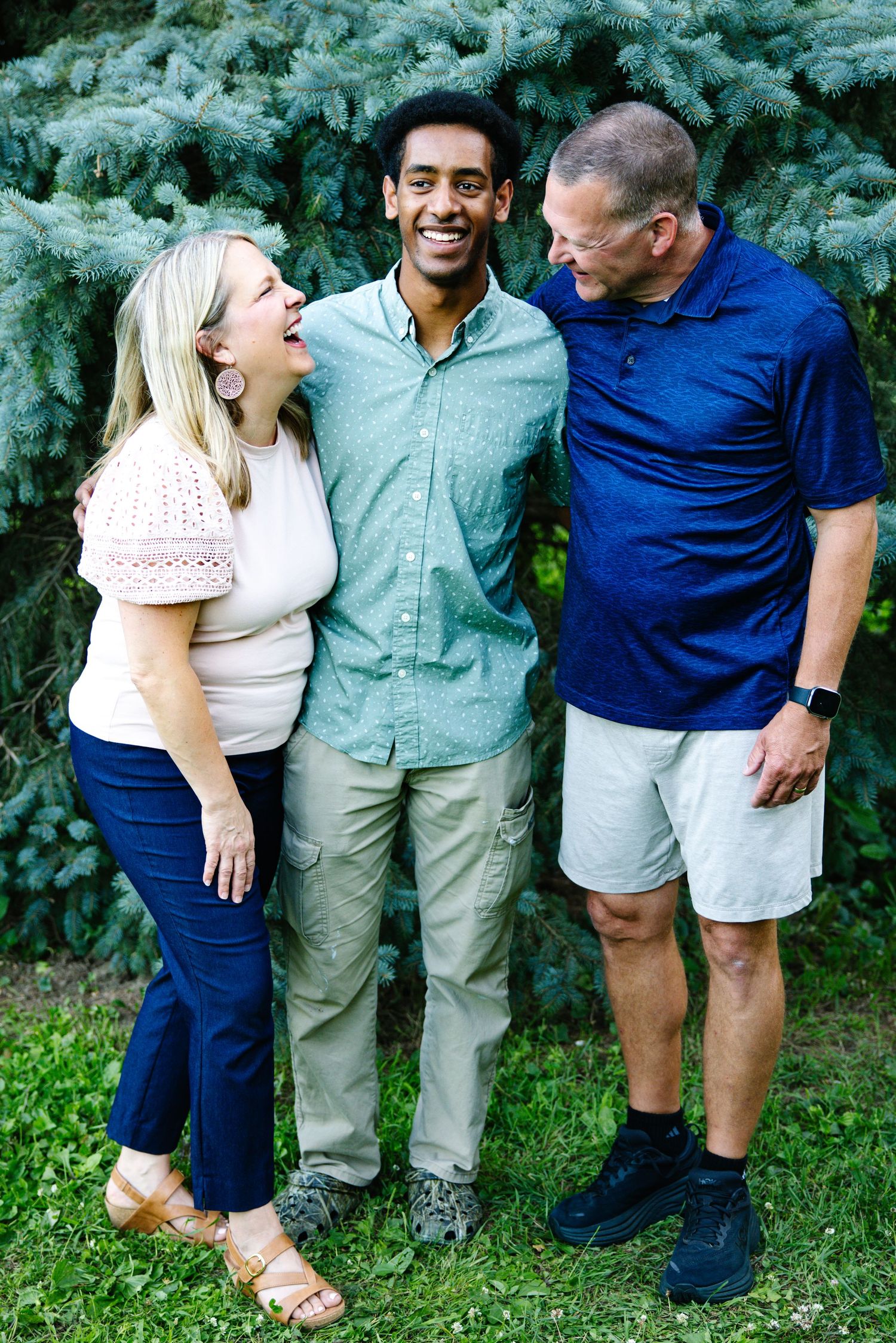 Three people laughing and smiling together outdoors in front of a blue spruce tree on a sunny day.