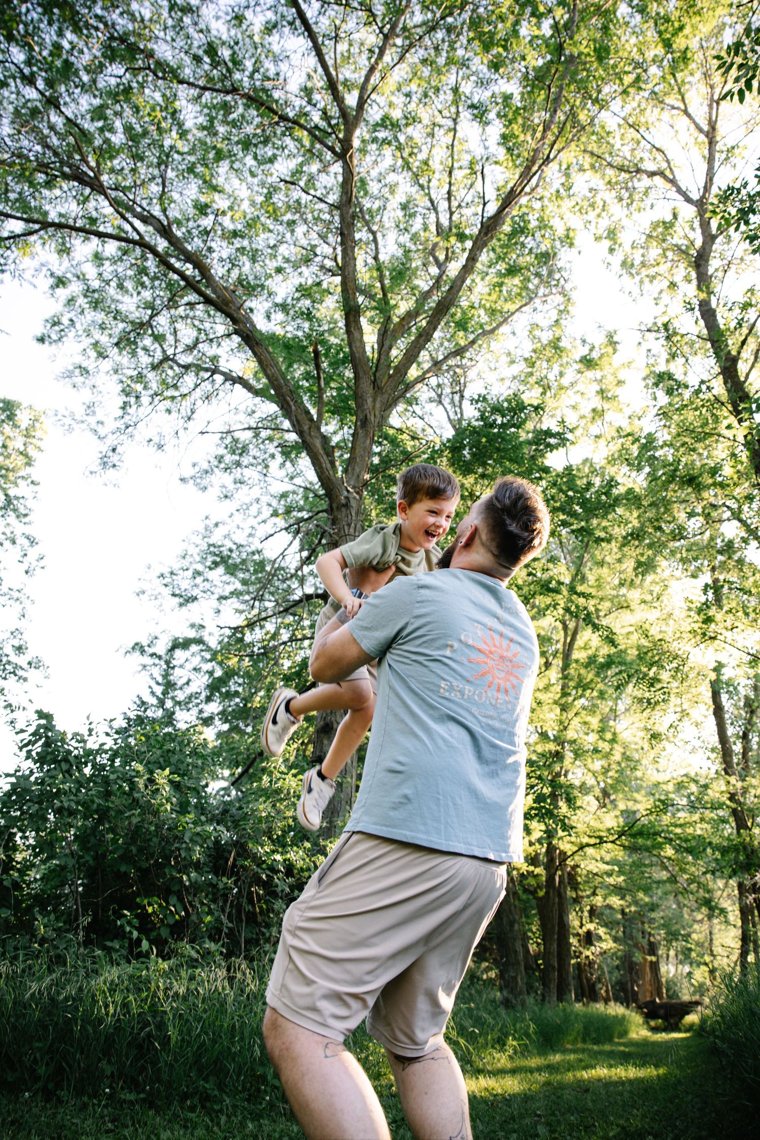 A father joyfully lifts his young son in a lush green forest, both smiling during a sunny outdoor play session.