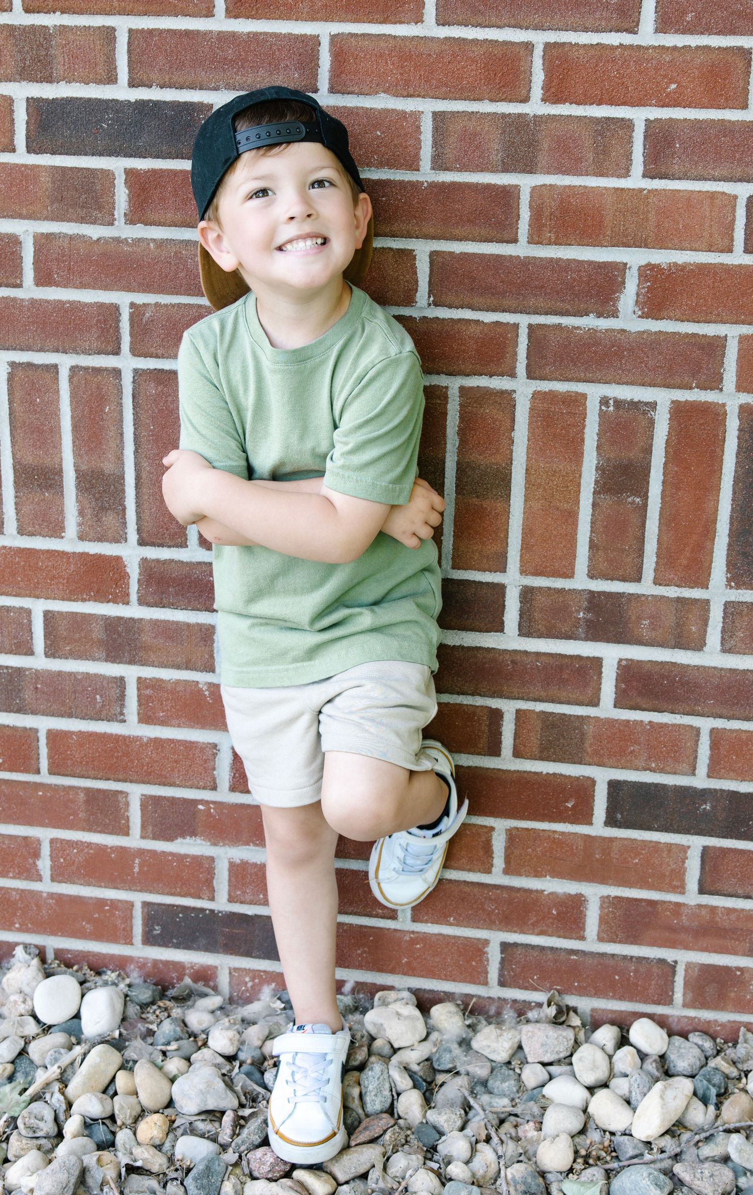 A smiling boy in a green tee, khaki shorts, and snapback hat leans against a brick wall.