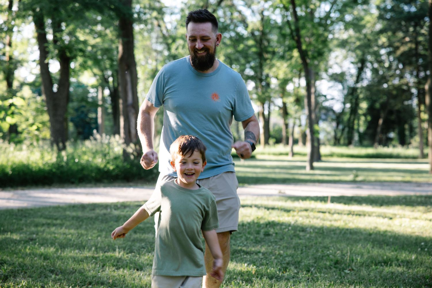 A bearded dad chases his laughing young son through a sunny green park on a warm summer day.