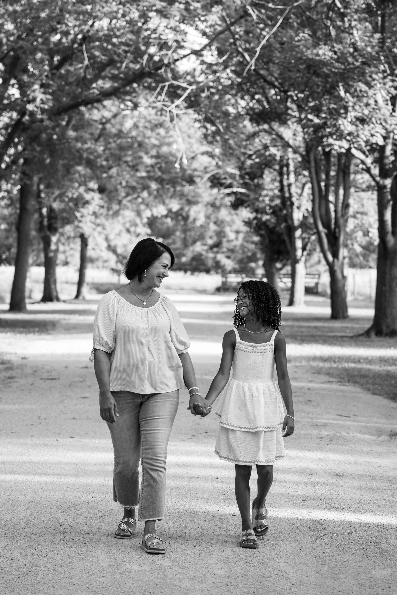 Black and white photo of a mother and daughter holding hands, walking together on a tree-lined path in a park.