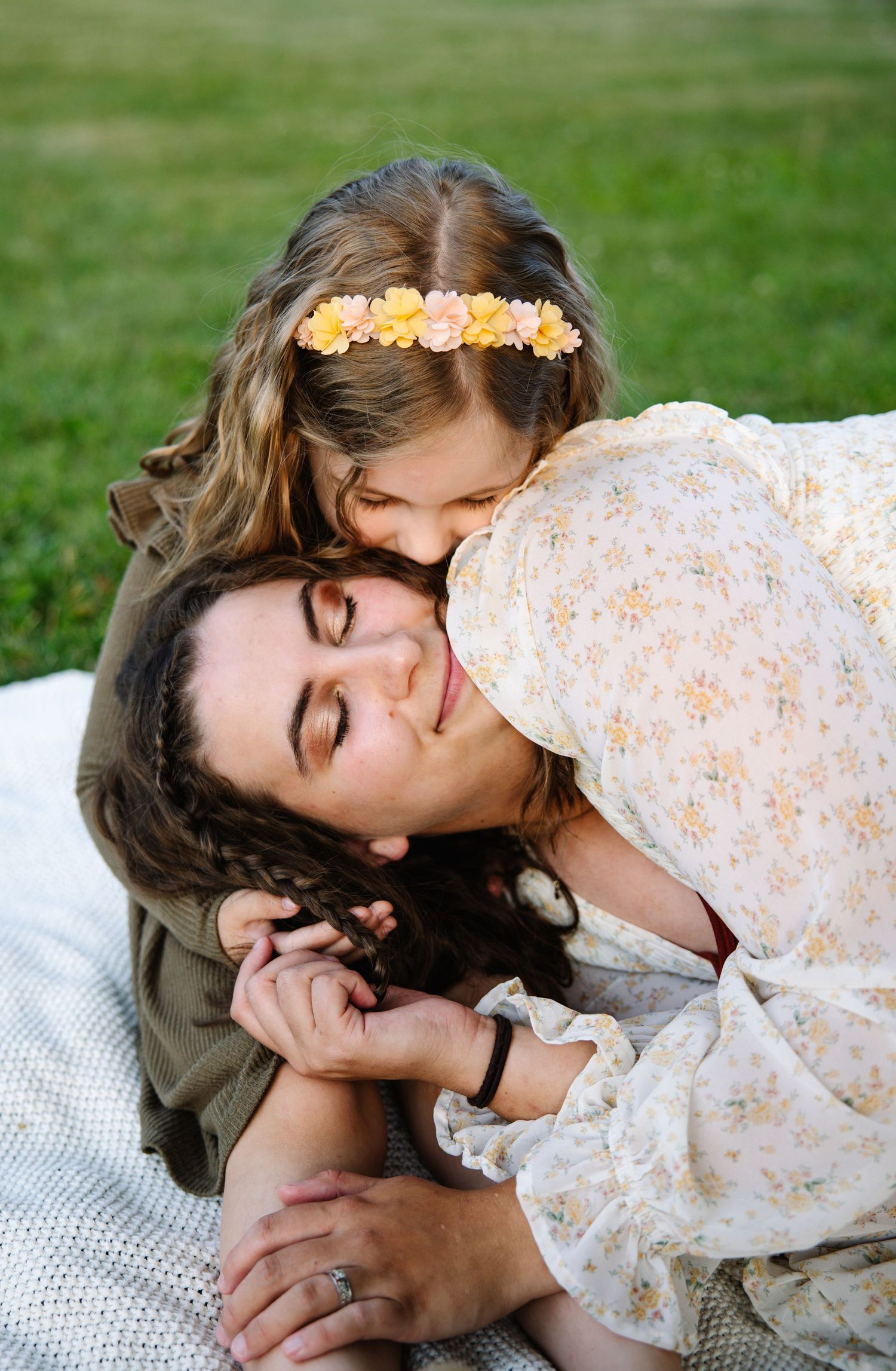A mother lies on a blanket outdoors as her young daughter wearing a floral crown leans down to kiss her cheek.
