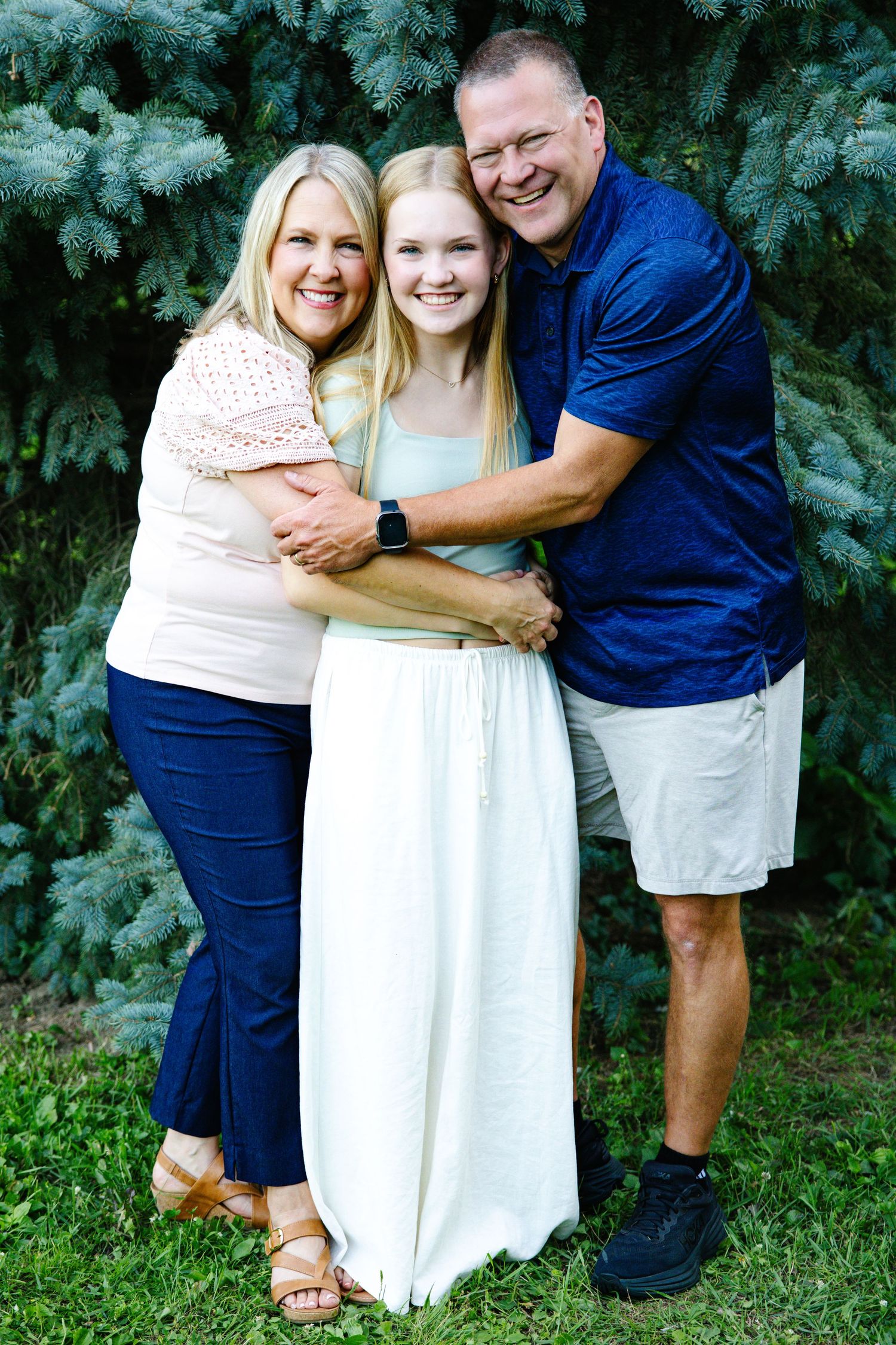 Happy family of three hugging outdoors near evergreen trees, smiling couple with teenage daughter in summer clothes.