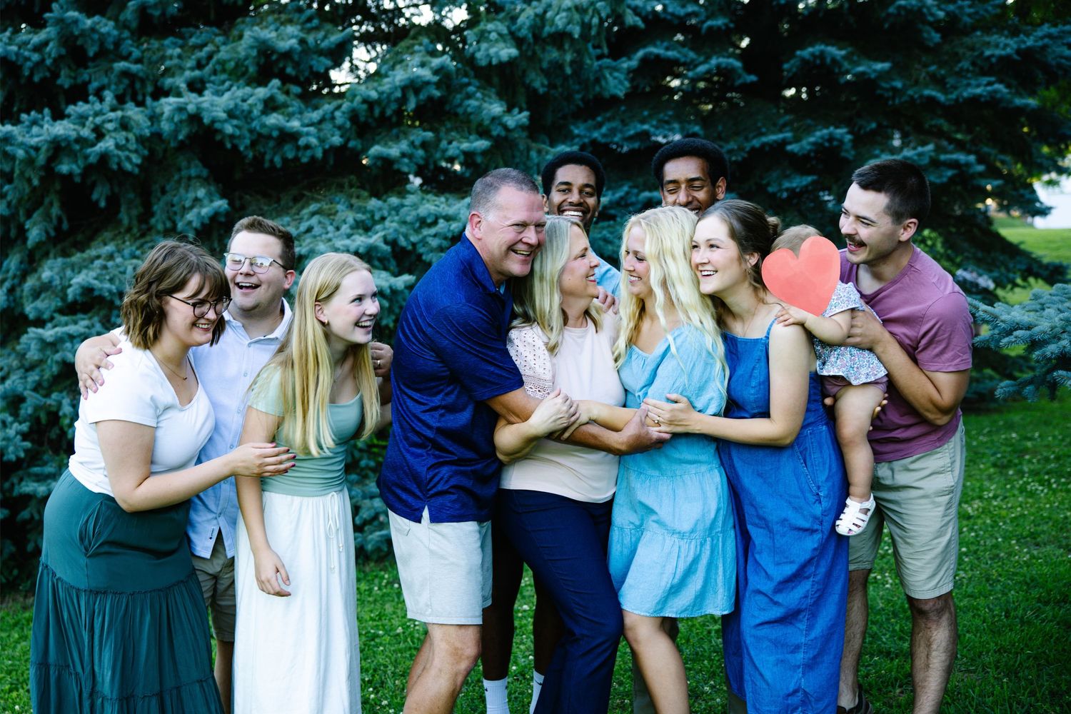 Large joyful family group laughing together outdoors near evergreen tree during casual photo session.