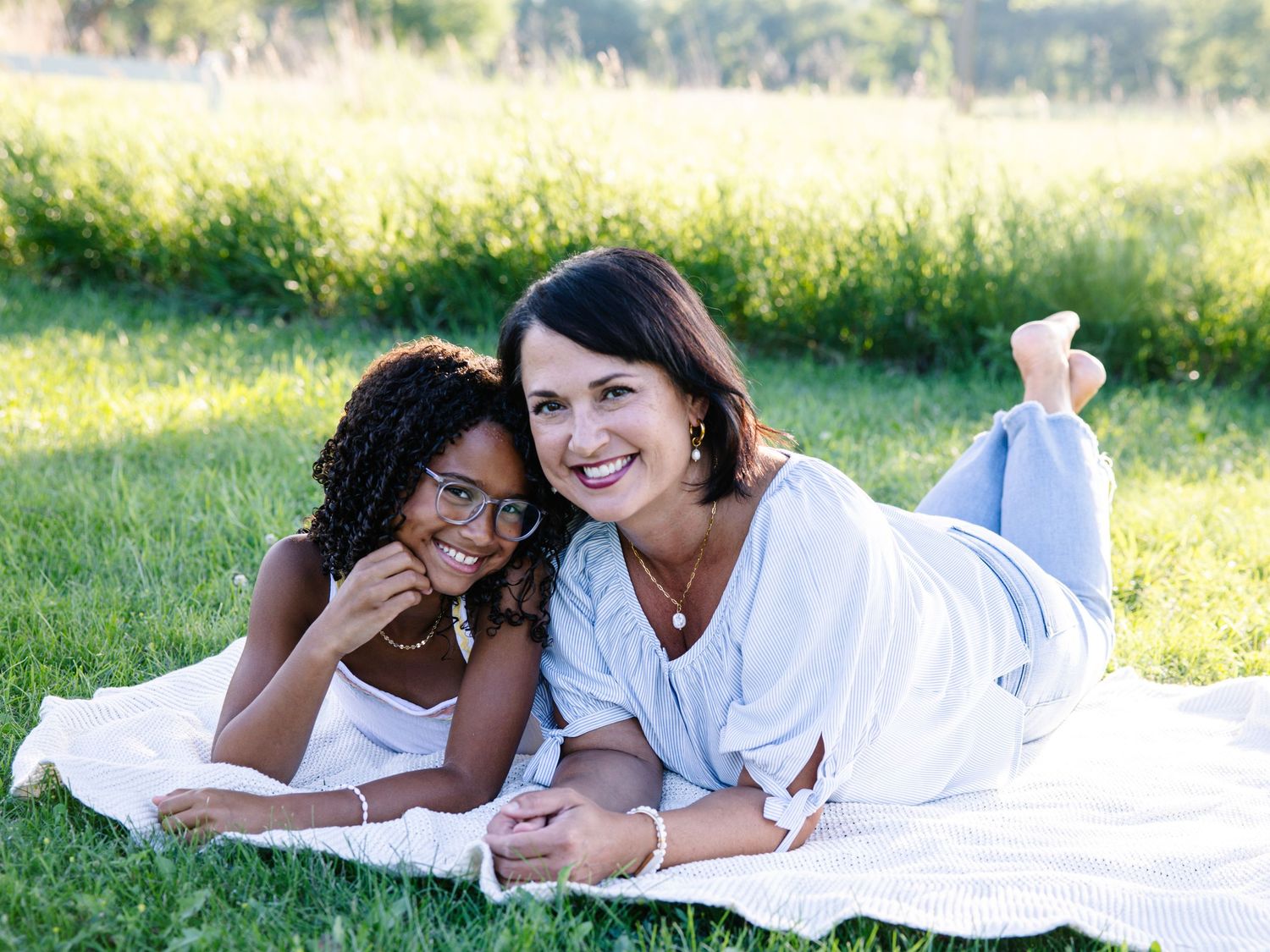 Two women smiling and lying on a blanket in a sunny green park, enjoying a warm outdoor day together.