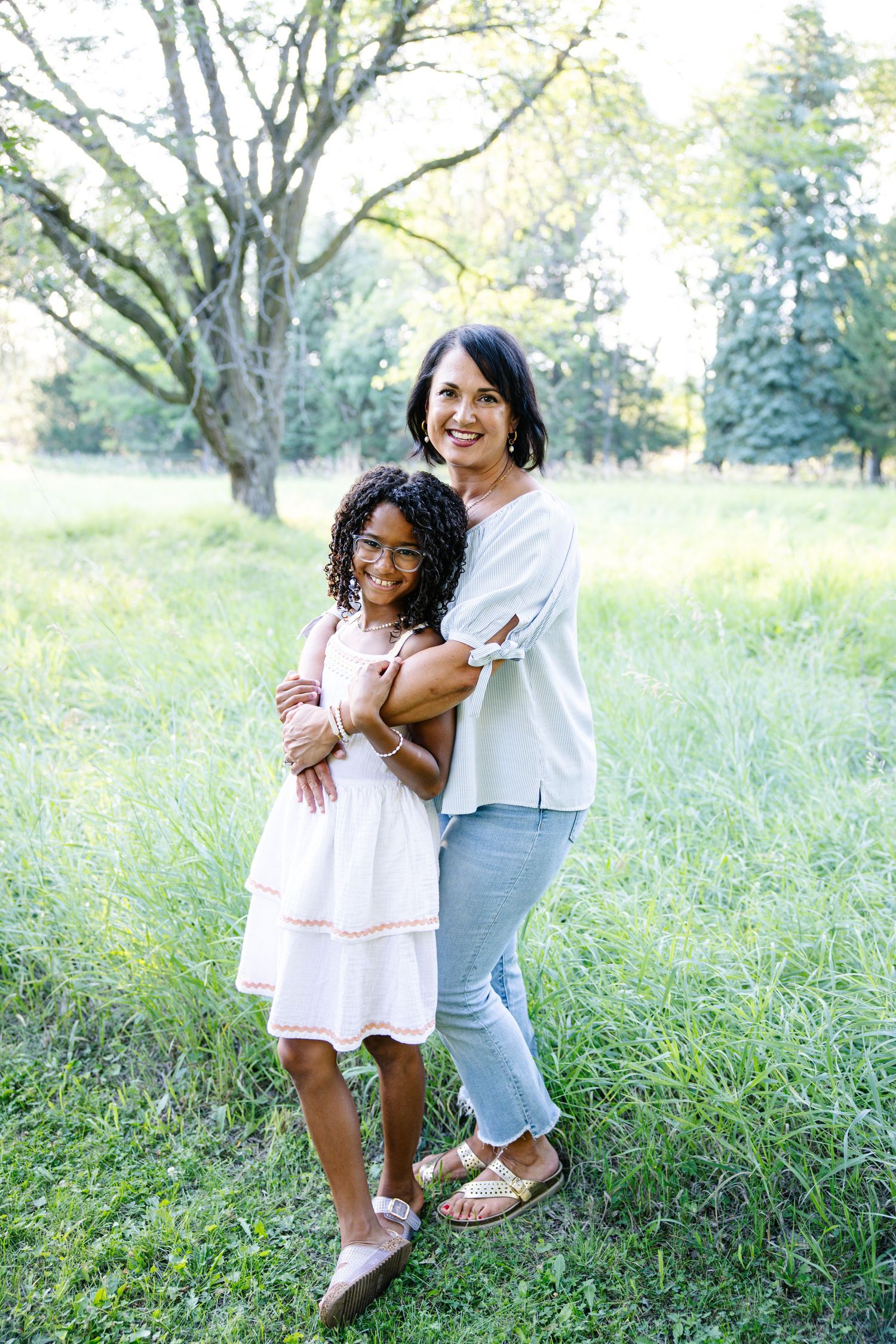 A woman and young girl in a white dress smile together in a lush green outdoor setting.
