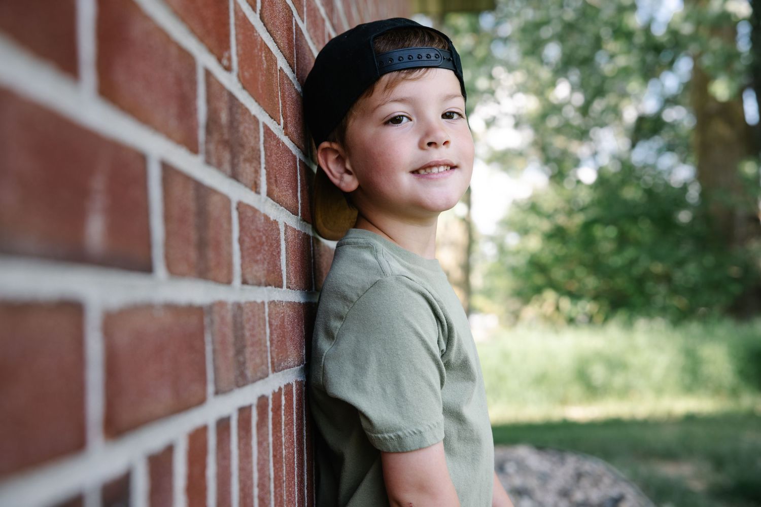 A cheerful boy in a green shirt and black snapback smiles while leaning on a brick wall.