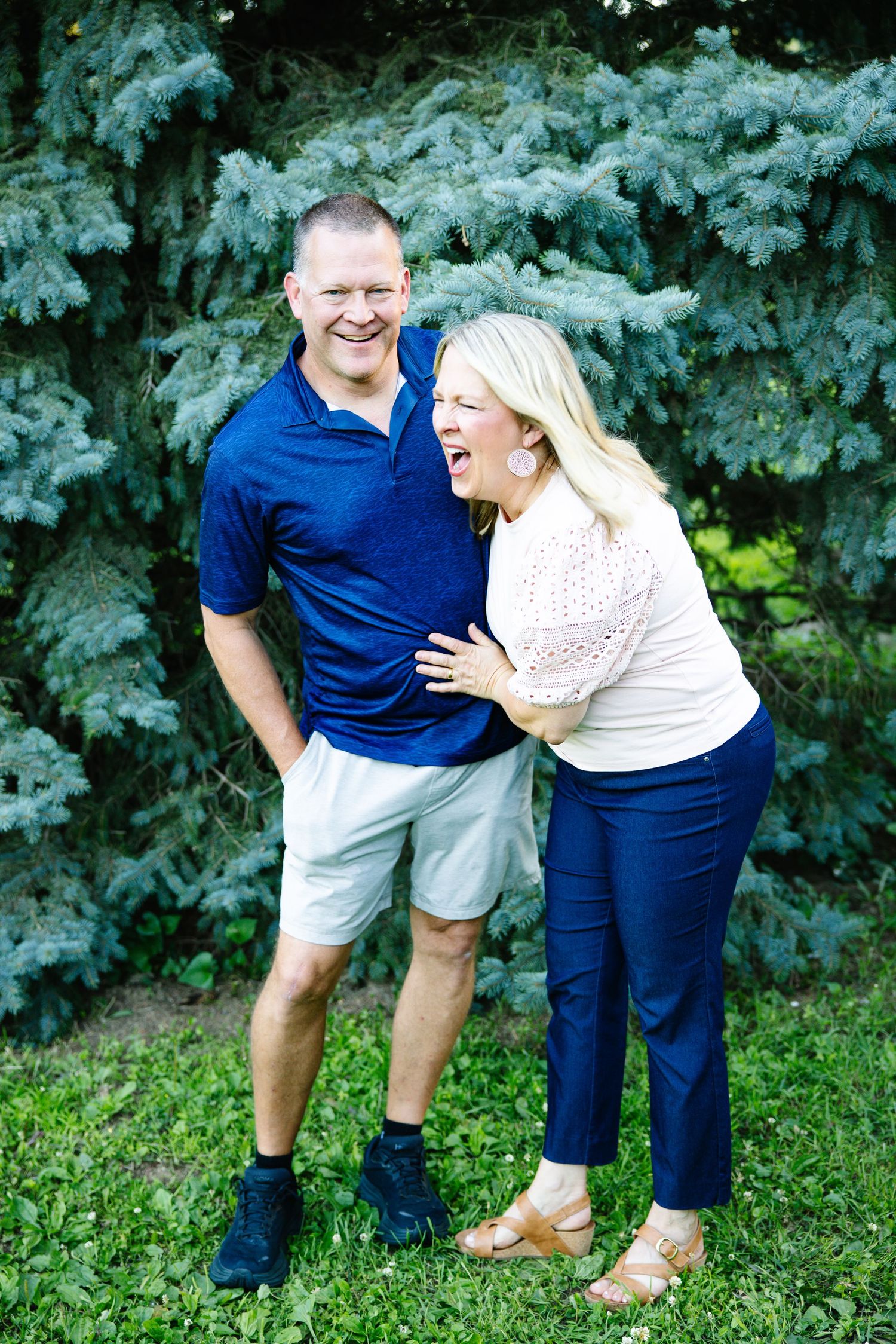 A laughing couple poses outdoors near evergreen trees, woman in pink top and navy pants, man in blue polo and white shorts.