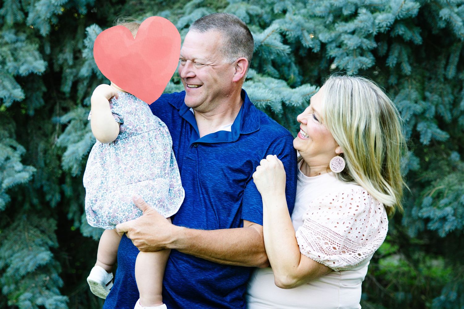 Happy couple smiling at each other while holding a young child outdoors near evergreen trees.