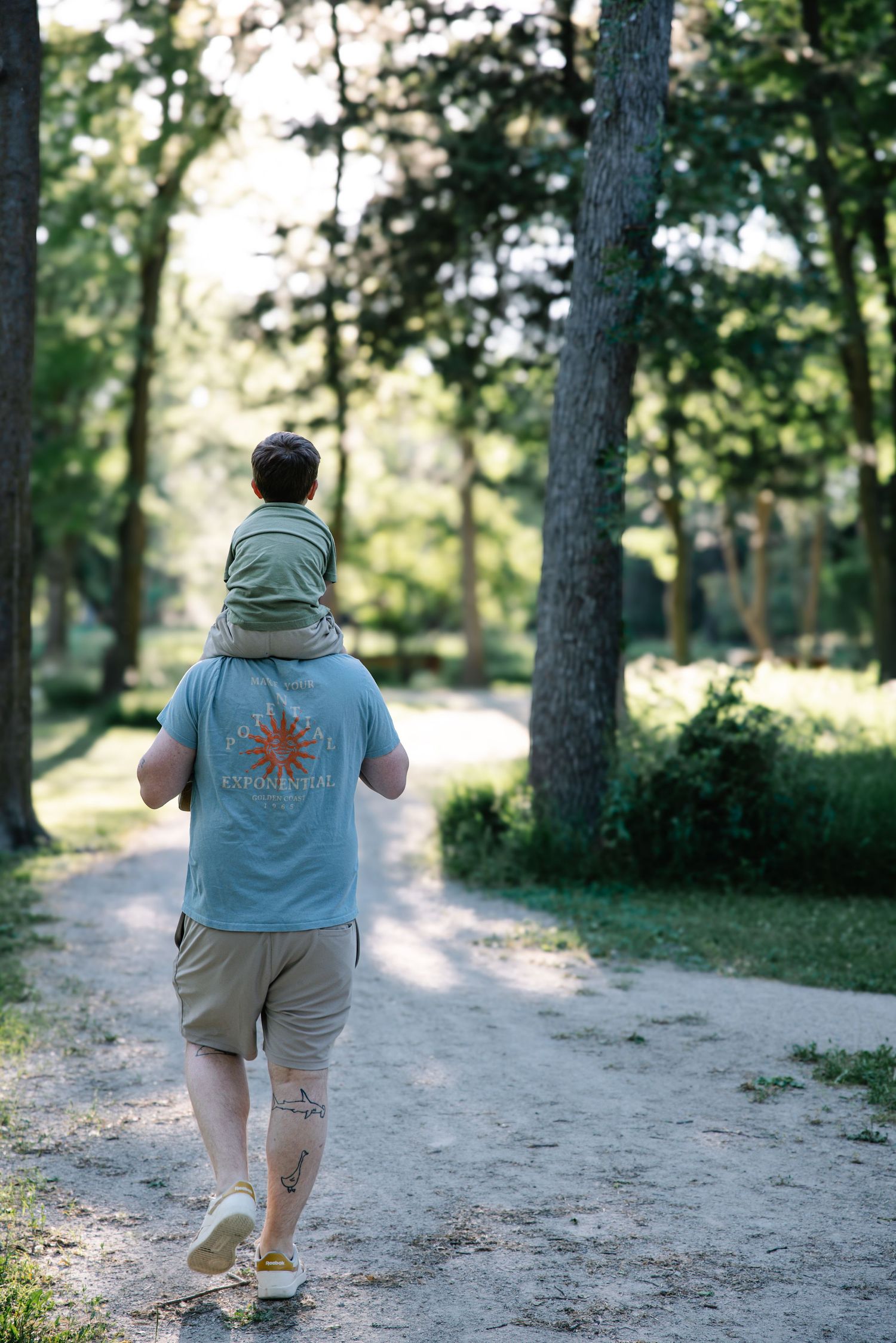 Father carrying young son on shoulders walking down a sunny park path surrounded by green trees.