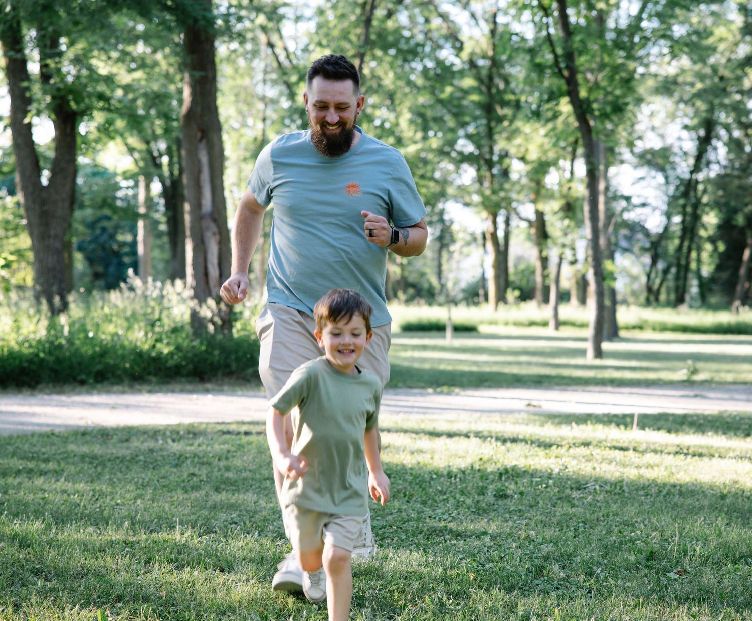 A laughing father chases his young son across a lush green park on a sunny day.
