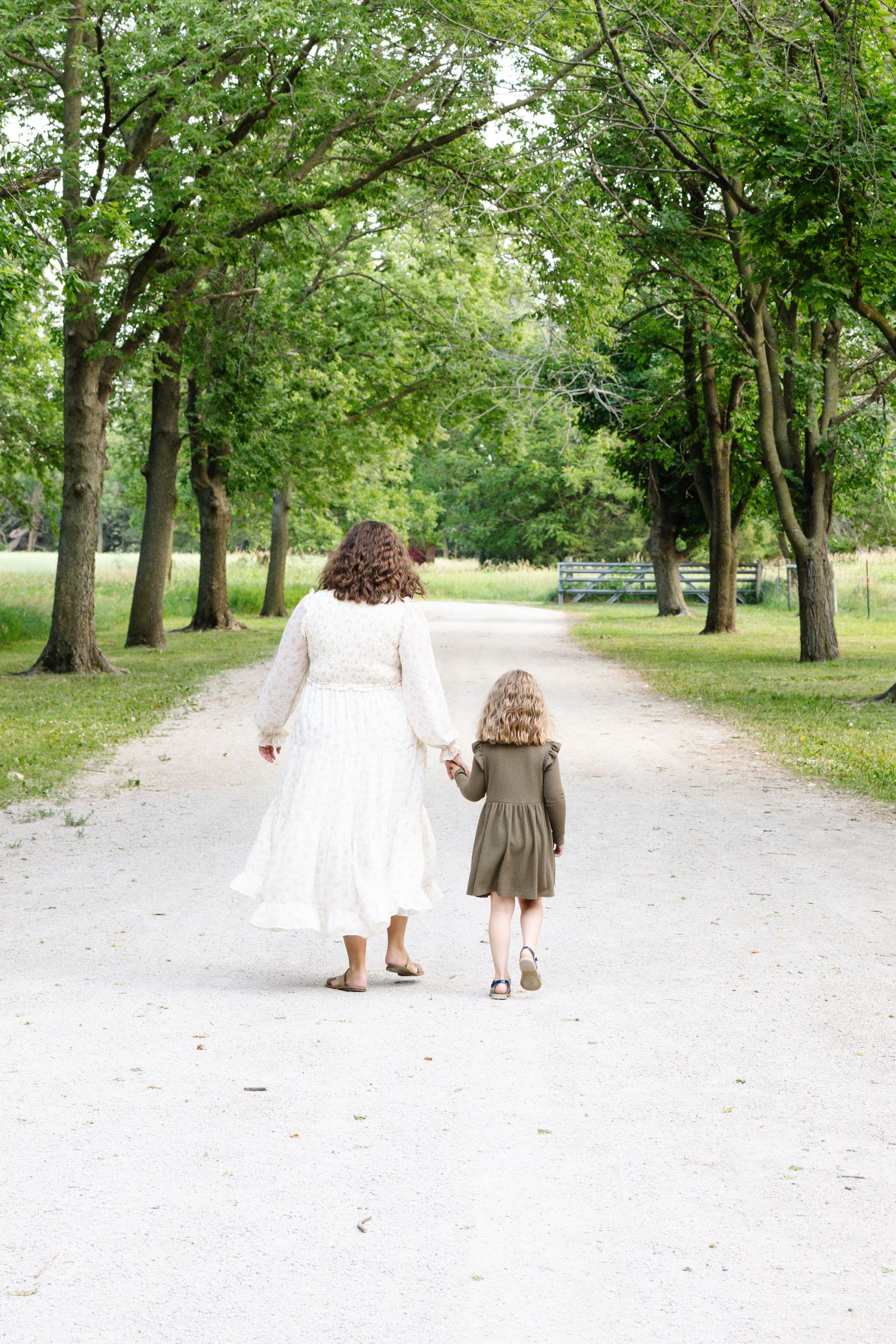 Mother and daughter walking hand in hand down a tree-lined path in a lush green park.