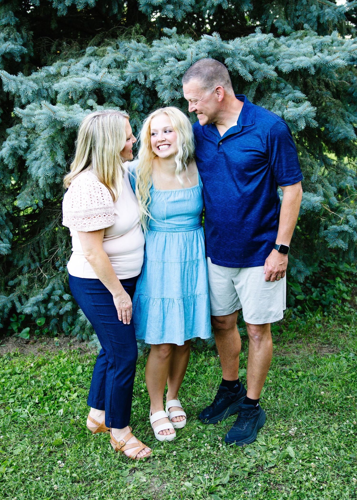 A smiling family of three poses outdoors near a blue spruce tree, with parents flanking their teenage daughter.