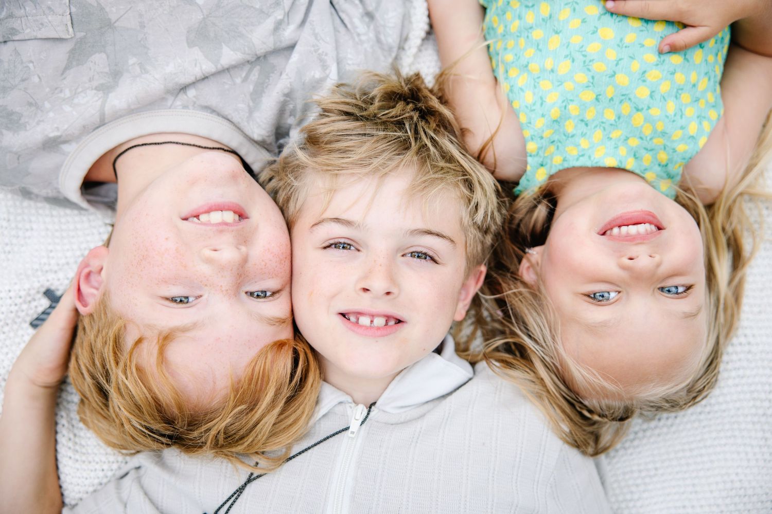 Three smiling blonde children lying together on a white blanket, looking up at the camera with joyful expressions.
