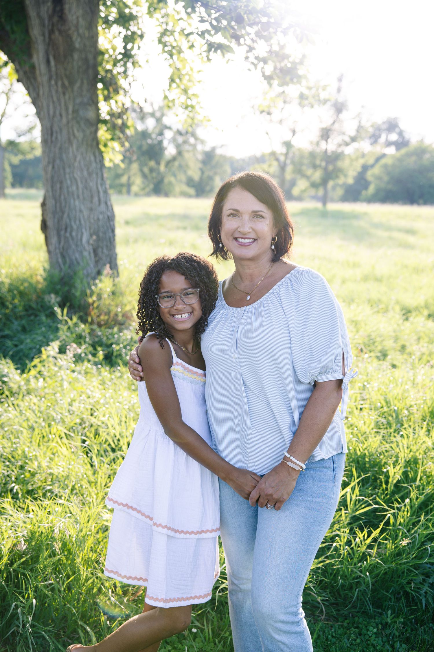 A woman and young girl smiling together outdoors in a sunny green park, wearing casual summer clothes.