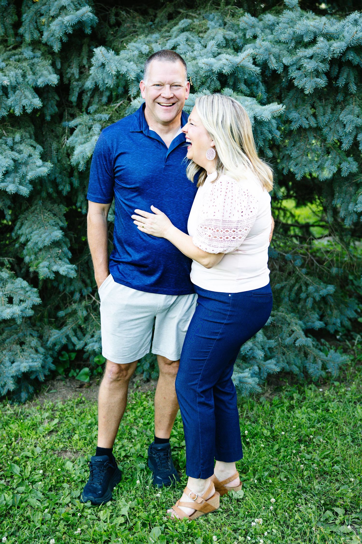 A smiling couple embraces outdoors in front of lush blue spruce trees, wearing casual summer attire.
