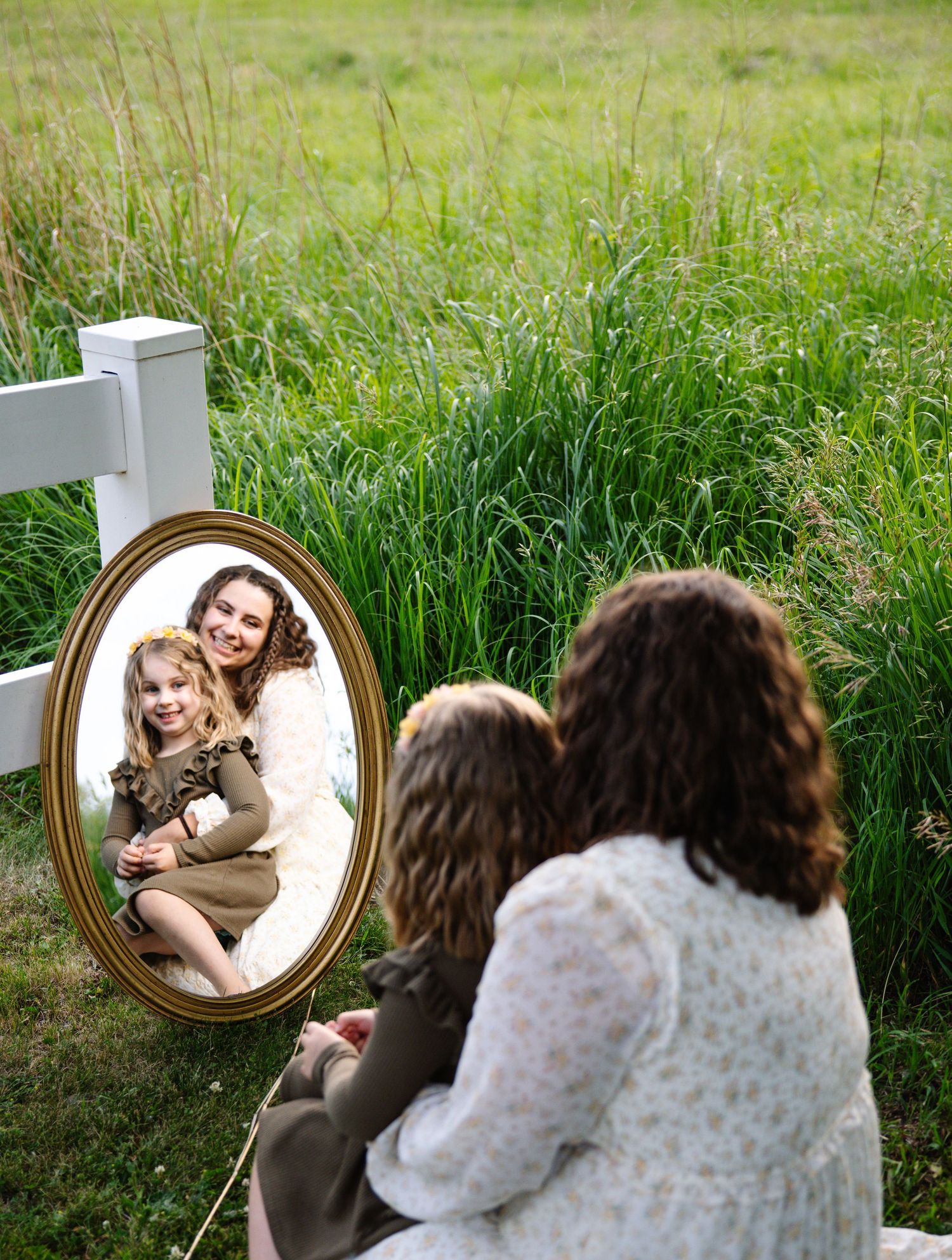 Mother and daughter sitting outdoors reflected in an oval gold-framed mirror in a green field.