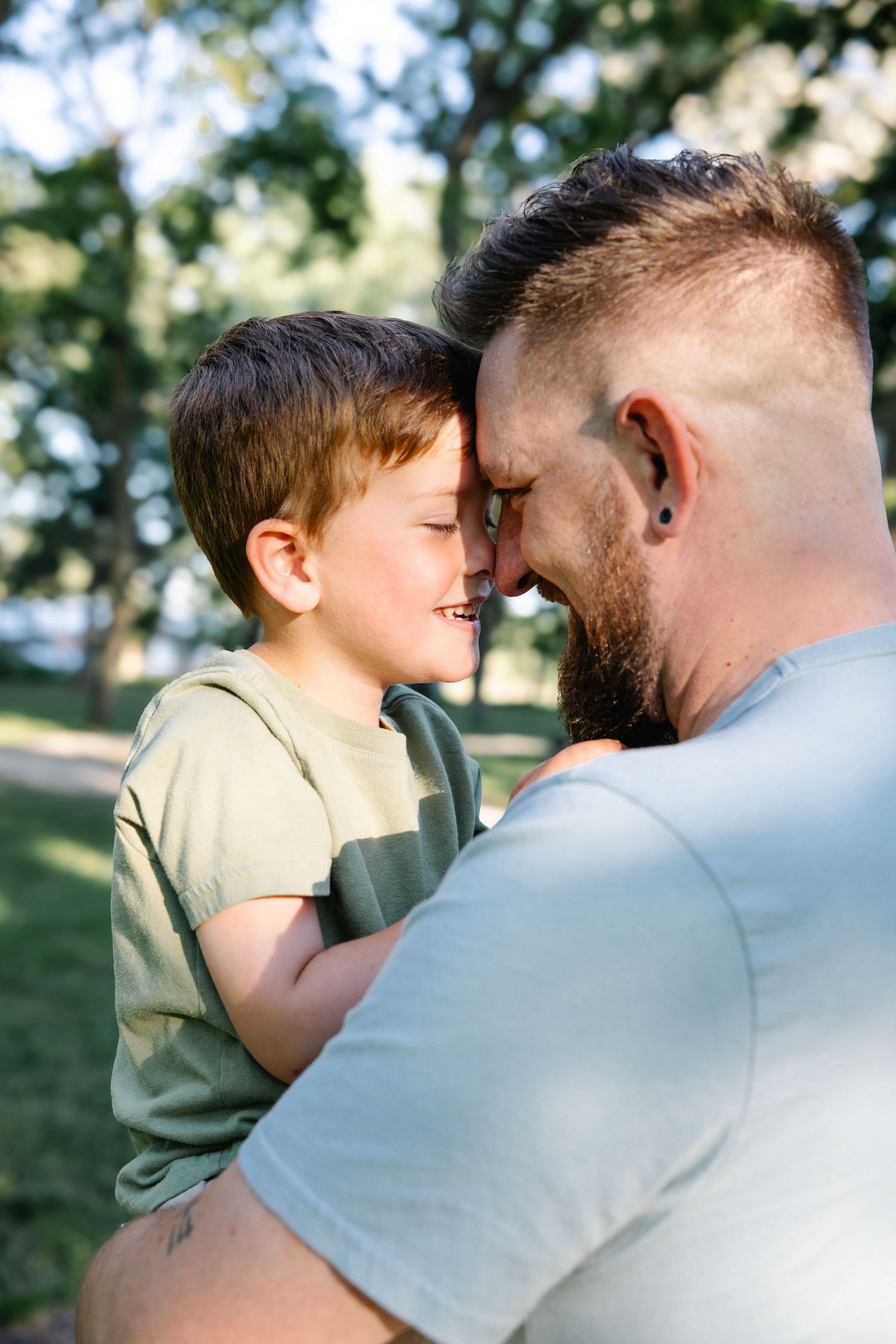 A father and young son touch foreheads and smile together in a sunny park, sharing a tender bonding moment.