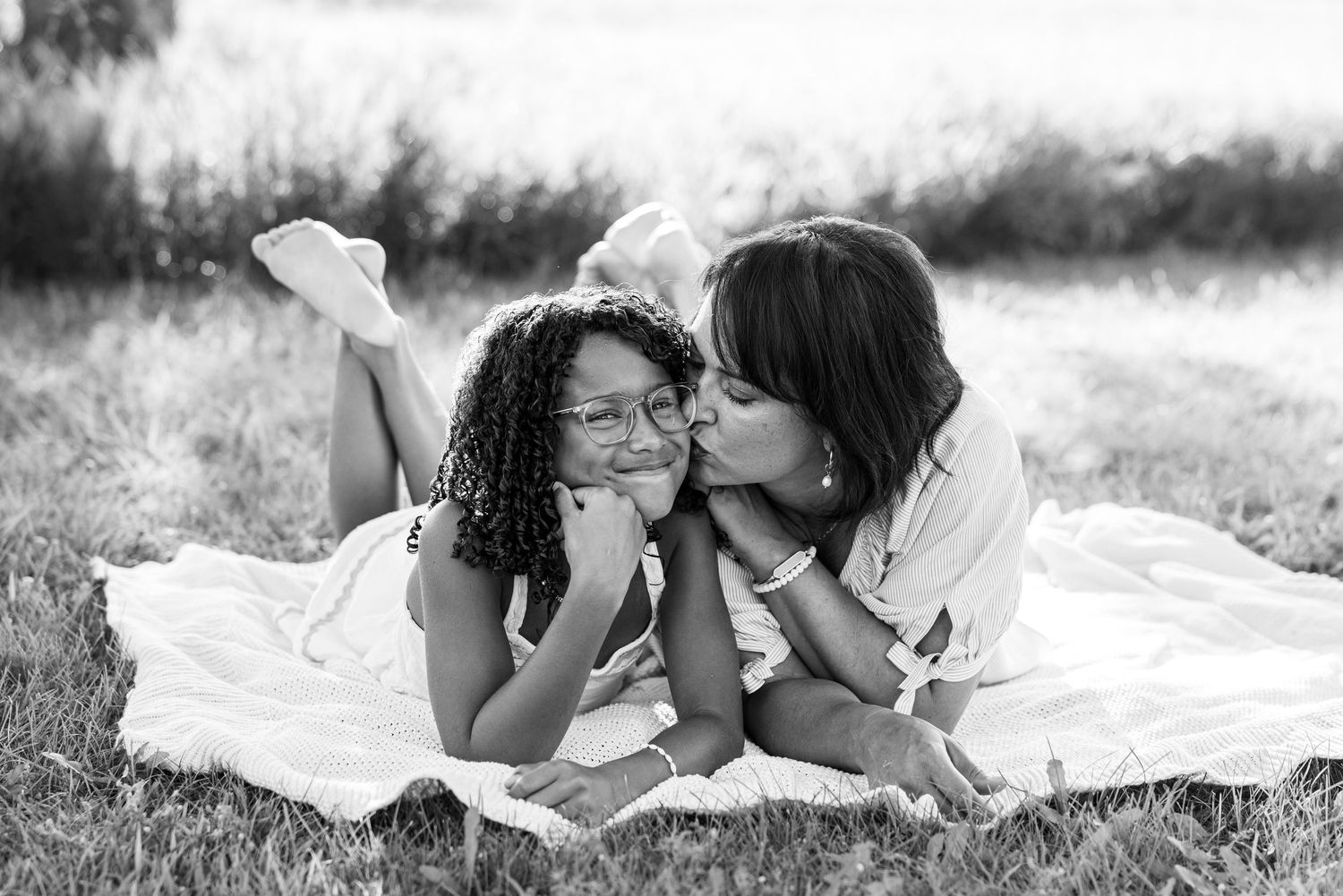 Black and white photo of a mother kissing her daughter on the cheek while lying on a blanket outdoors.