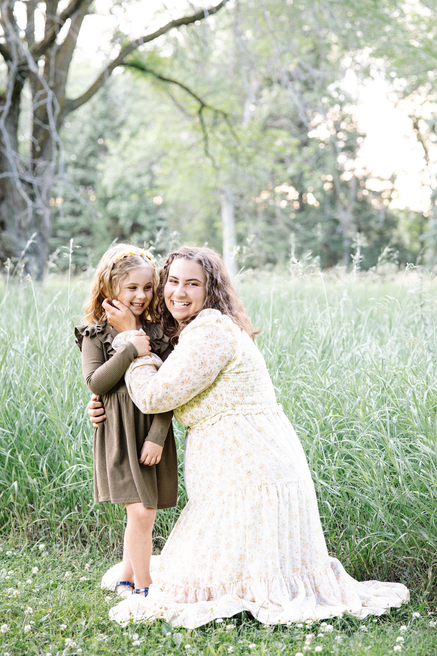 A smiling mother in a floral dress hugs her young daughter in a green field.