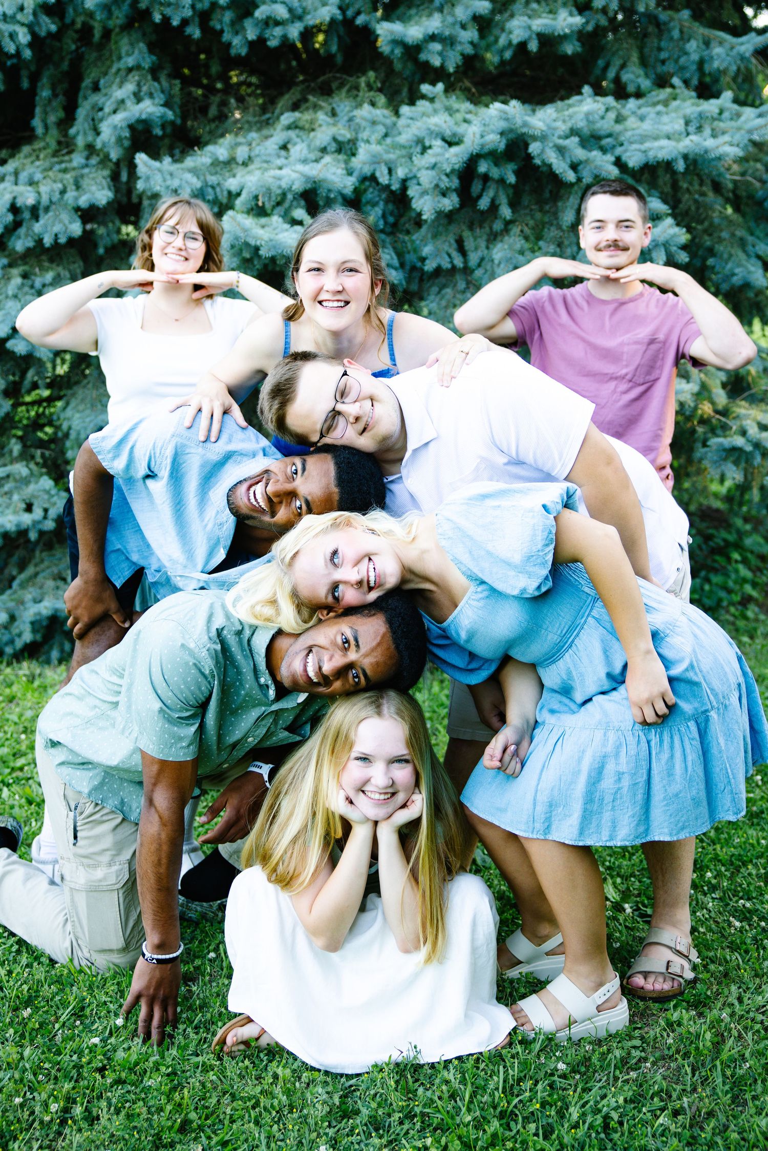 A diverse group of young adults posing playfully in a pyramid formation on green grass outdoors.