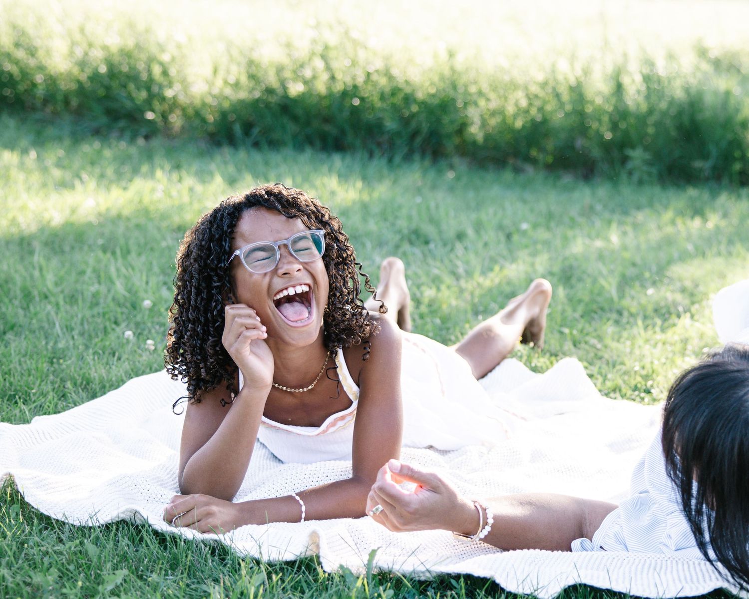 A young woman with curly hair and glasses laughs joyfully while lying on a blanket in a sunny green park.