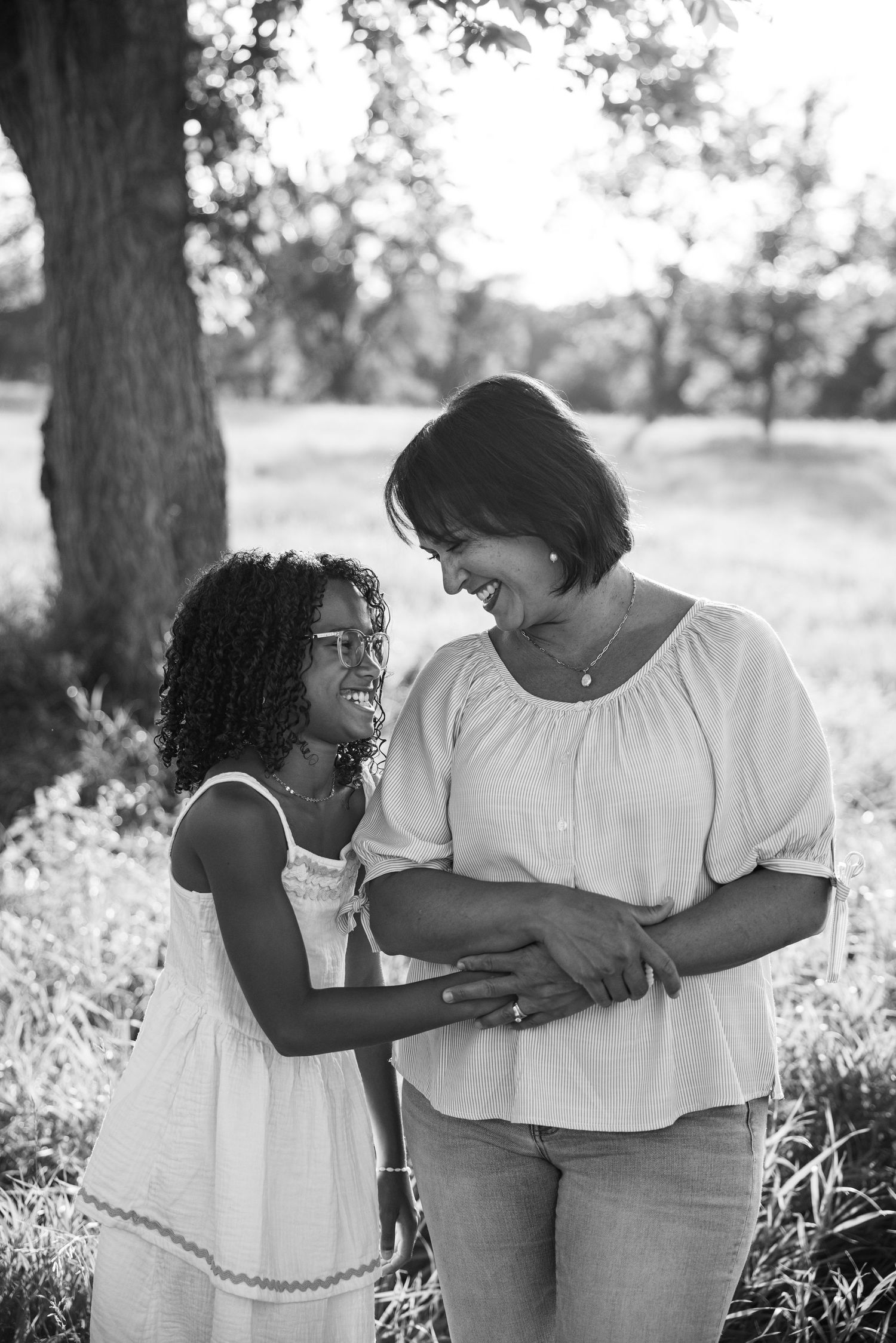 Black and white photo of a woman and young girl smiling at each other outdoors in a park setting.