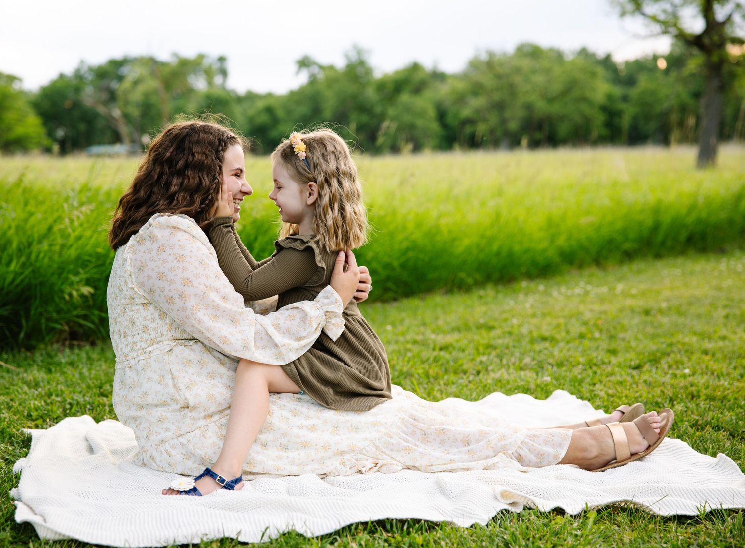 A mother and young daughter sit together on a white blanket in a lush green park, sharing a tender moment.