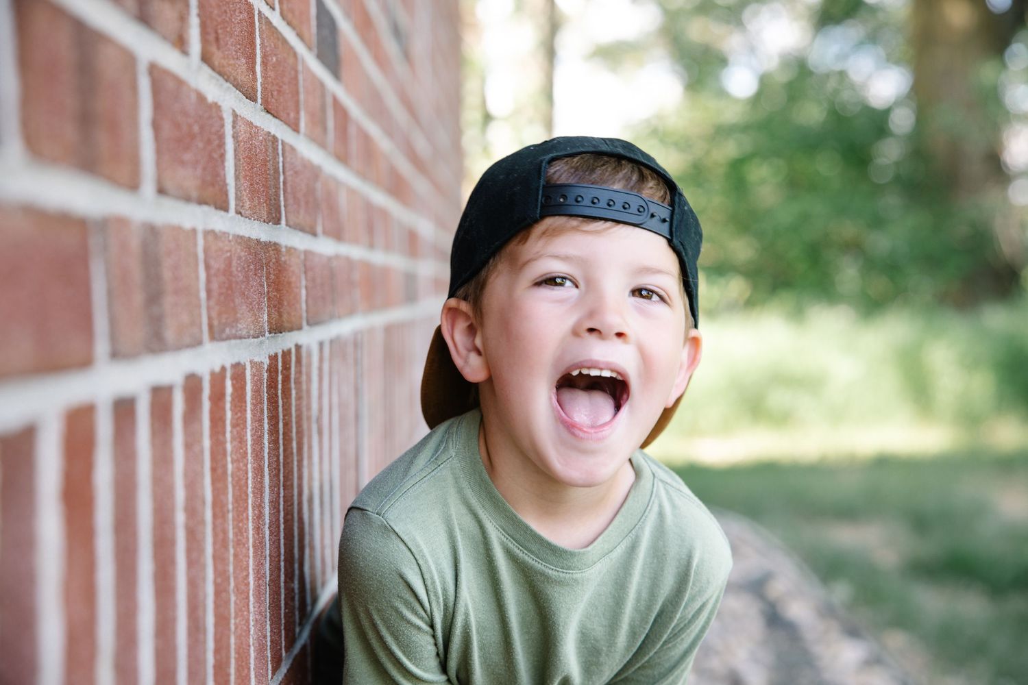 Happy young boy in a backward black cap laughs against a brick wall outdoors.