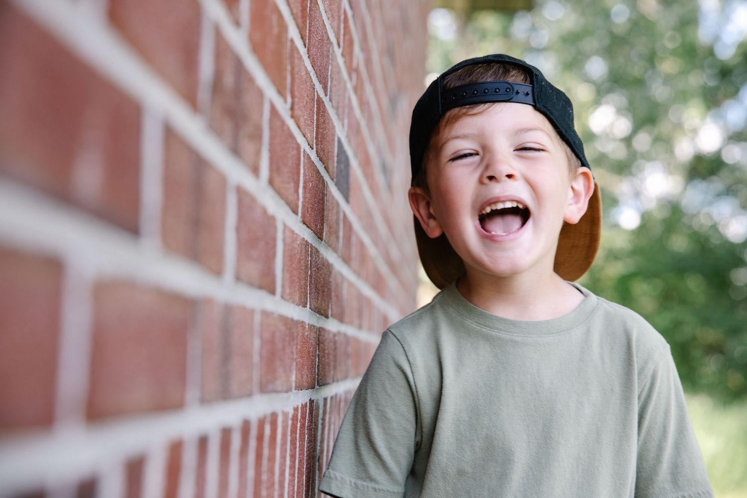 Happy young boy laughing against a brick wall, wearing a backwards cap and green t-shirt outdoors.