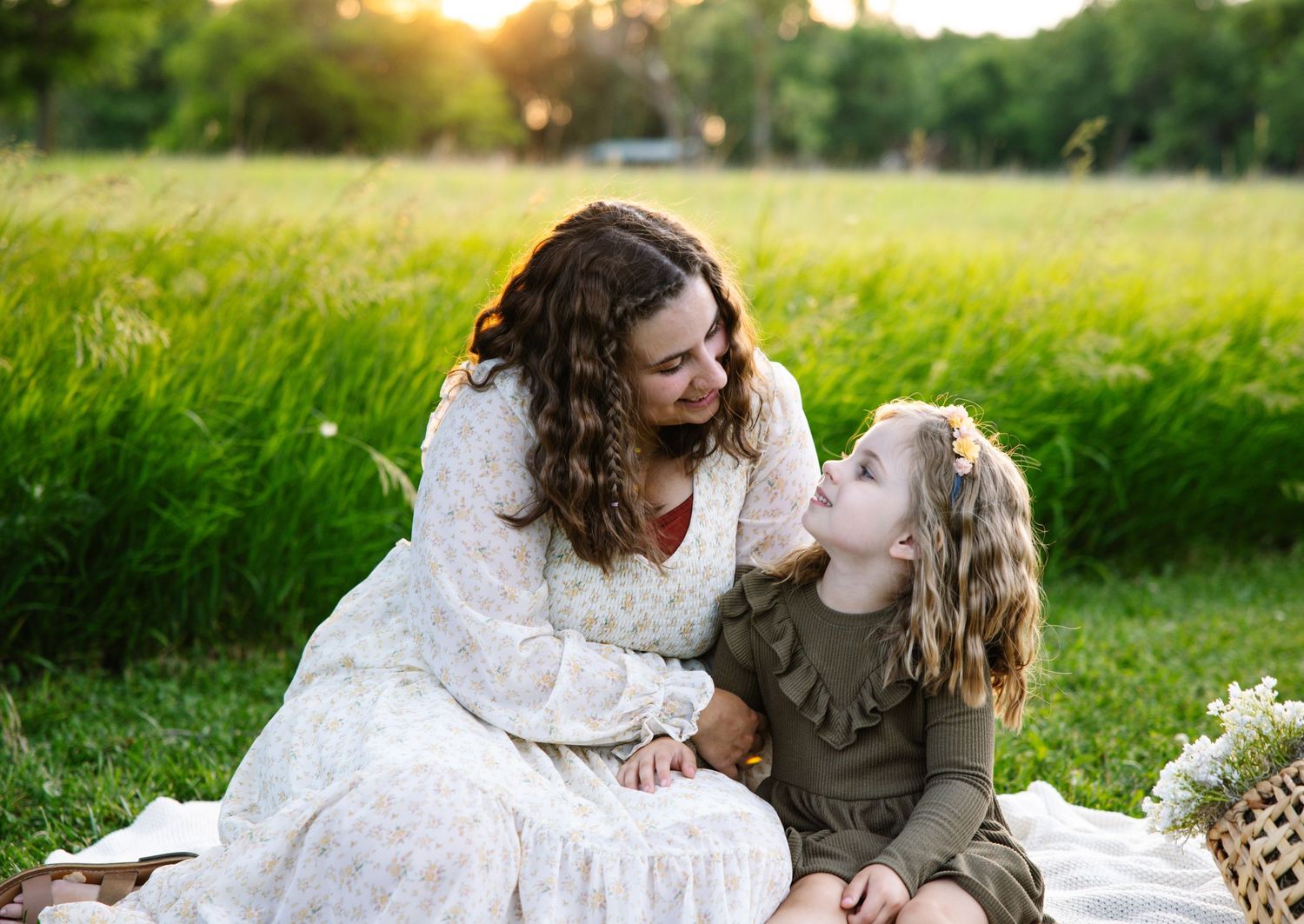 A mother and daughter share a tender moment sitting on a blanket in a sunlit green field.