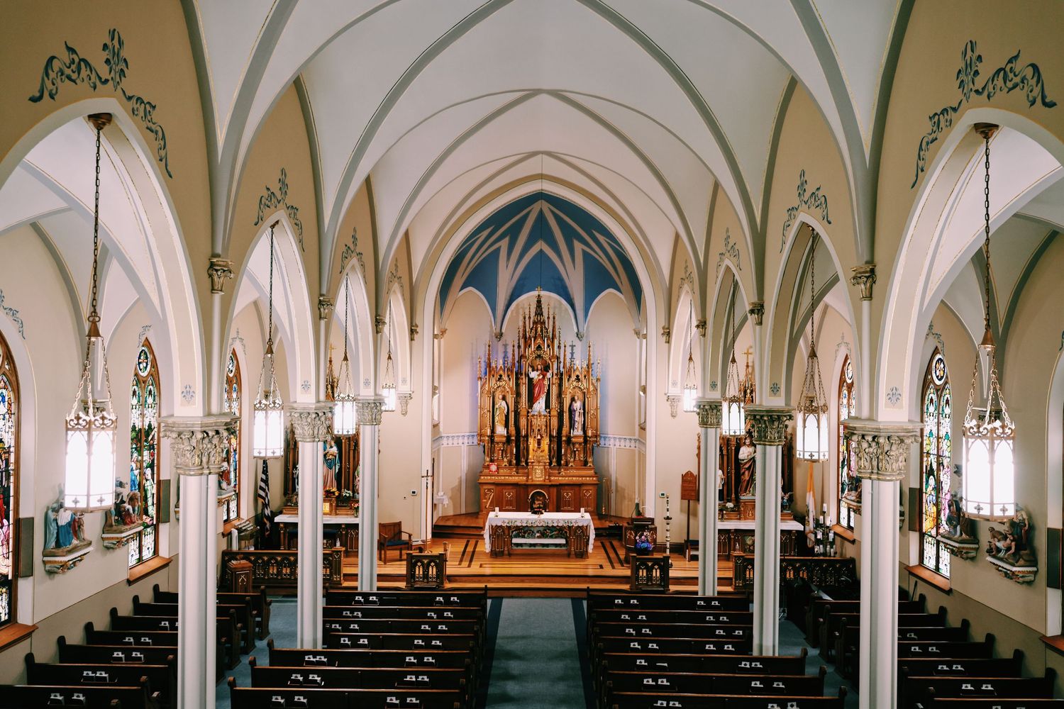 Interior of a grand Catholic church featuring white Gothic arches, wooden pews, stained glass windows, and ornate golden altar.