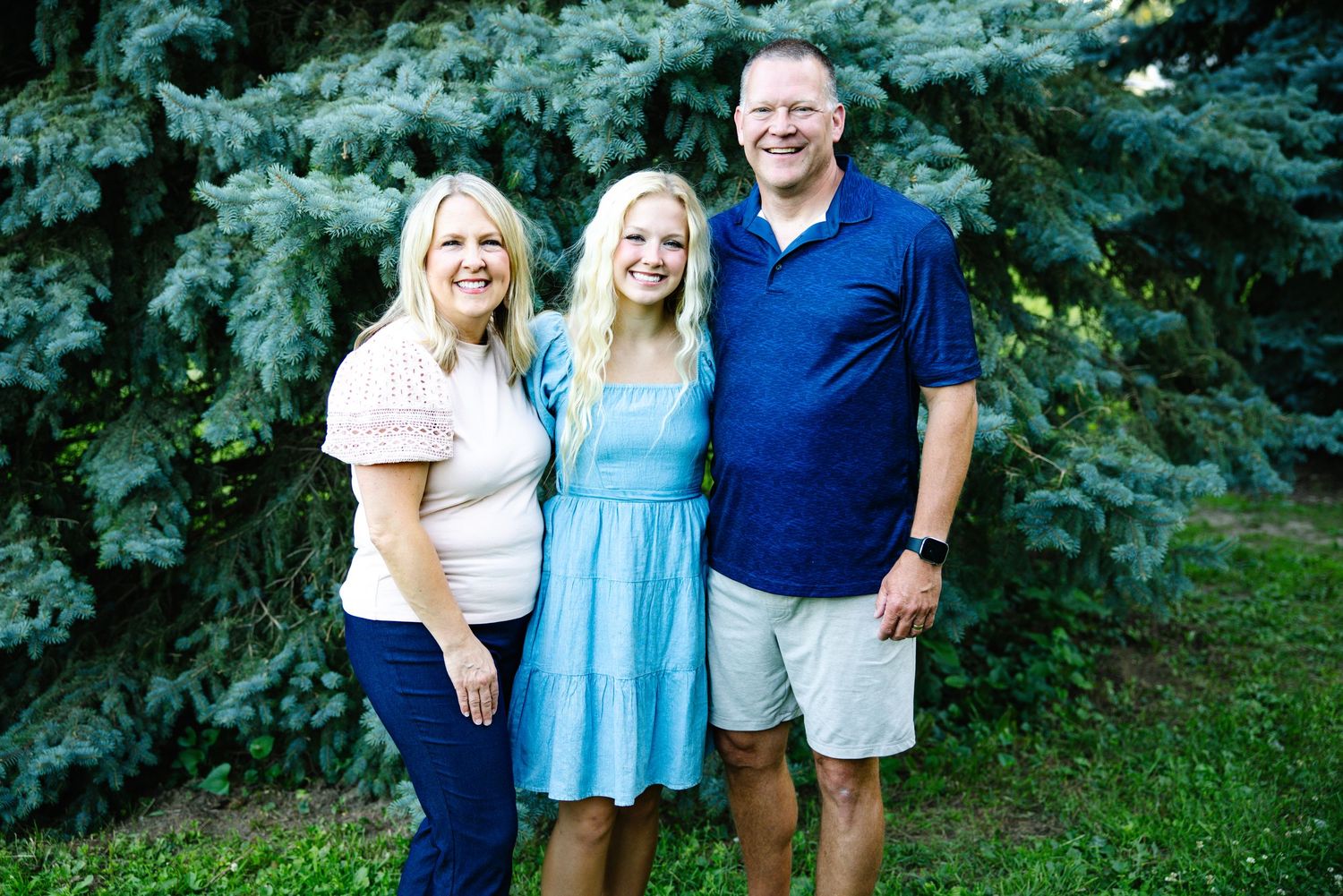 A smiling family of three poses outdoors in front of a lush green spruce tree on a sunny day.
