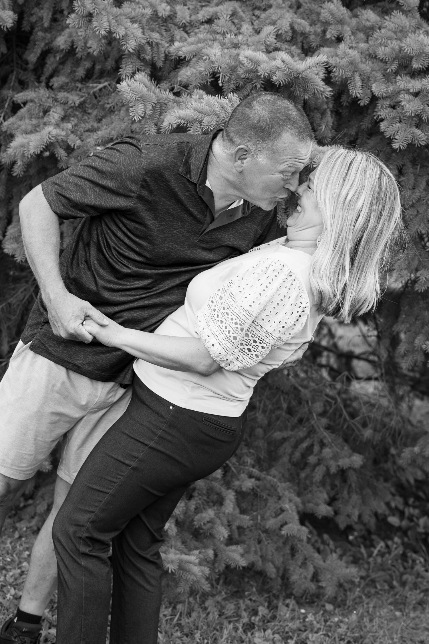 Black and white photo of a couple sharing a romantic dip kiss outdoors near evergreen trees.